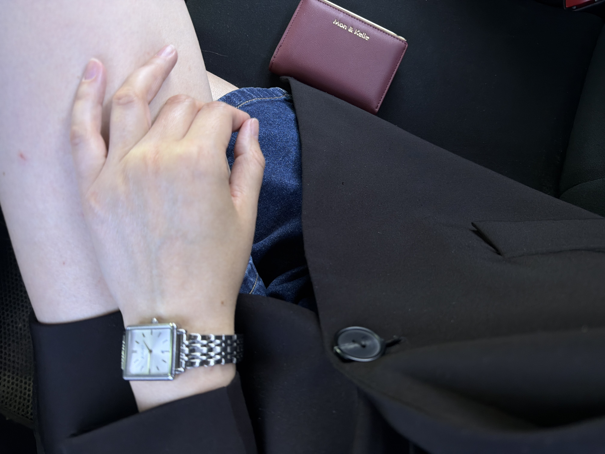 This is a close-up, over-the-shoulder shot of a person sitting in a car. The person is wearing a black blazer over blue jeans and a silver watch. Their hands are clasped in their lap, and a small burgundy wallet with gold lettering that reads "Mon & Kelle" is resting on the seat next to them. The interior of the car appears to be dark. The image does not contain any specific visual cues to determine the exact location within Chaikovsky, Russia.