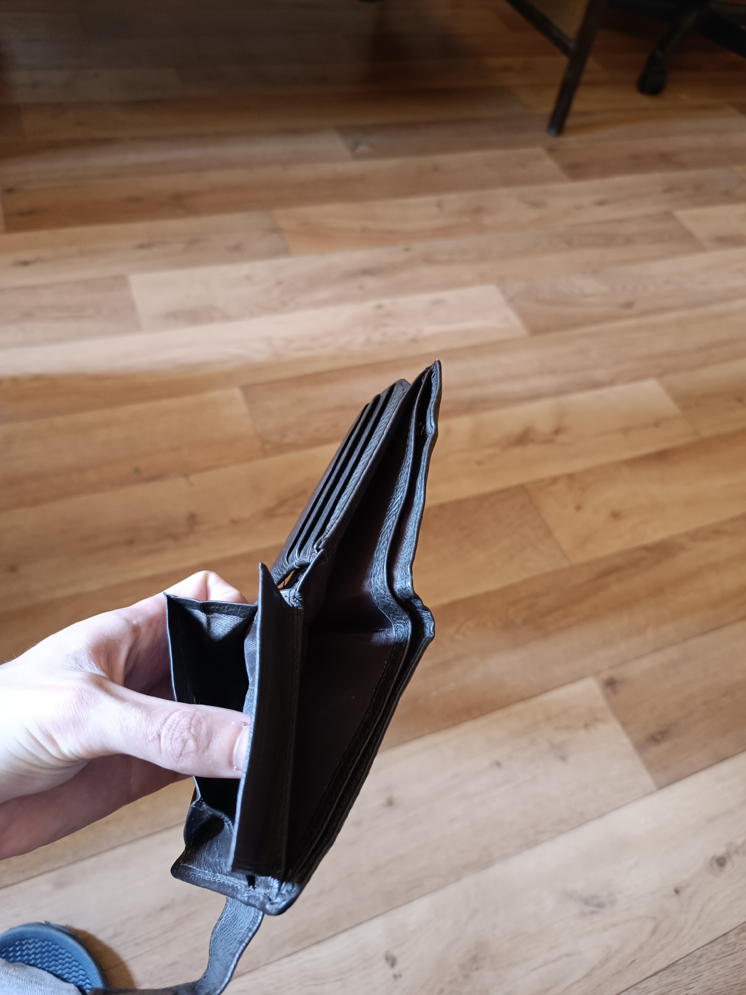 A partially opened, empty, dark leather wallet is held in a person's hand. The wallet is positioned against a background of light brown, wood-patterned flooring. A dark chair leg is visible in the upper right corner of the image. Part of a gray shoe sole is present in the lower left corner. There is no indication of Sofia, Bulgaria in the image.