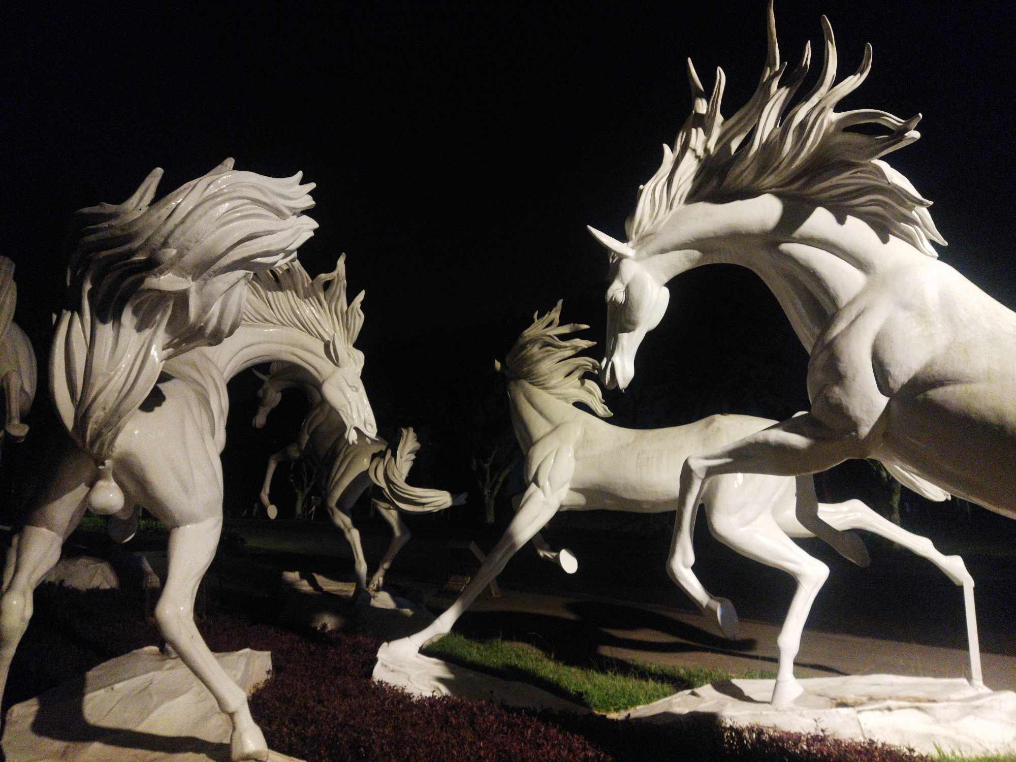 This image depicts a night-time scene featuring a collection of white horse sculptures arranged in a garden or park-like setting. The sculptures are detailed and appear to be in motion, with flowing manes and muscular bodies. The primary focus is on the large, dynamic sculptures in the foreground and mid-ground. The background is dark, suggesting it's nighttime, with some faint hints of trees or foliage. The ground beneath the sculptures is covered in a combination of low-lying plants and what appears to be a dark, textured mulch. The sculptures are illuminated, casting shadows and highlighting their forms. This artwork is located at the Lawang Sewu heritage building in Semarang, Indonesia.