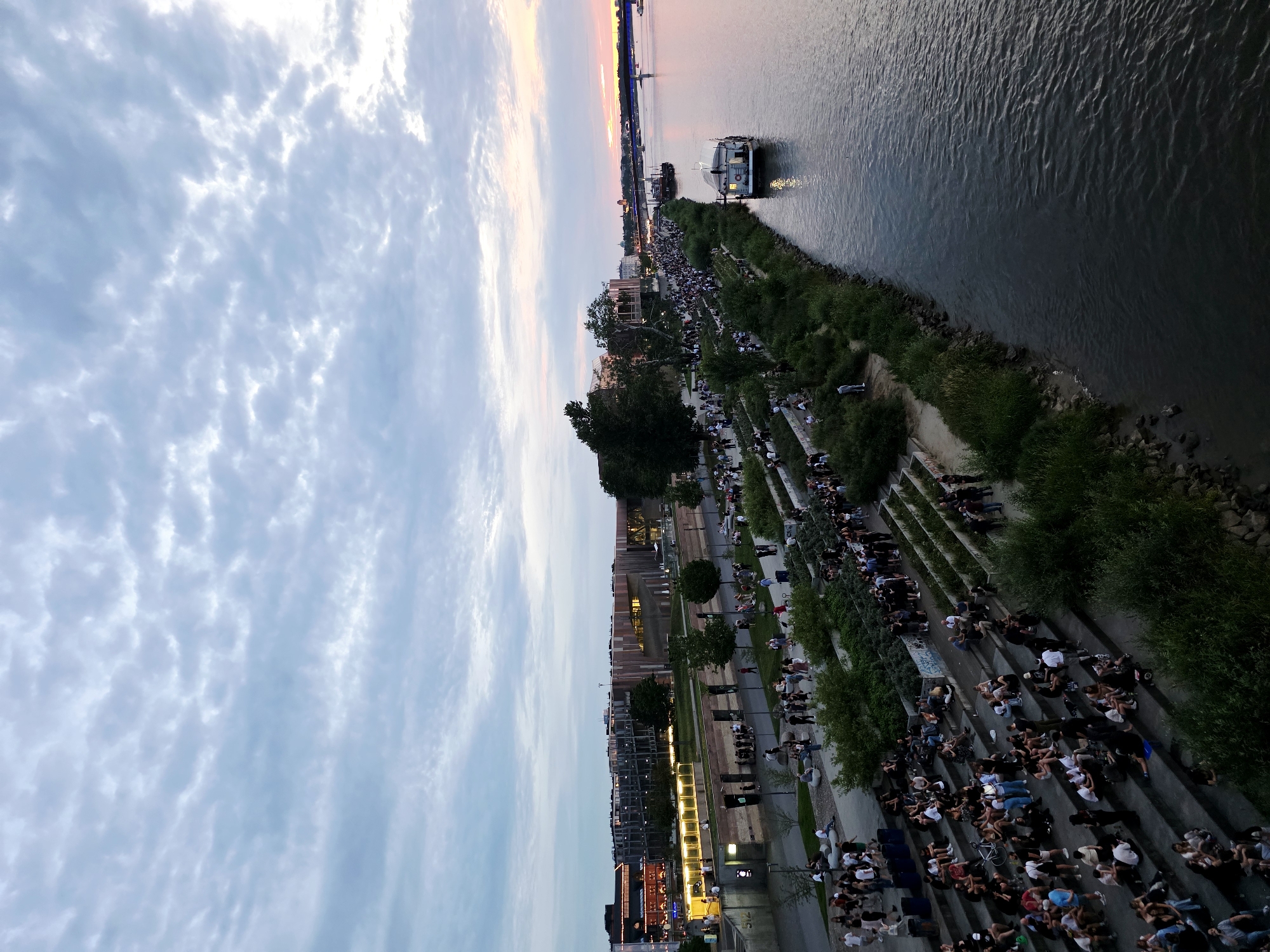 This is an aerial, wide-angle, high-angle shot of a bustling urban riverside promenade in Warsaw, Poland, during dusk. The Vistula River occupies the right two-thirds of the frame, its surface rippling with a dark, muted hue. A white houseboat with a single window is docked on the river, close to the shore. Further out in the river, a larger vessel, possibly a ferry, is visible, along with a distant bridge spanning the water.
The left side of the image depicts a tiered, terraced walkway packed with a large crowd of people. They are seated on the steps, socializing, and enjoying the evening. The terraces are interspersed with lush green vegetation and planters. Behind the terraces, a pedestrian walkway runs parallel to the river, also populated with many individuals. Beyond this walkway, a modern urban cityscape is visible. Buildings with contemporary architecture, featuring glass facades and intricate patterns, are illuminated, suggesting commercial or residential use. Streetlights line the pathways, casting a warm glow.
The sky above is a canvas of dramatic clouds, with patches of lighter hues indicating the fading sunlight of sunset. The overall atmosphere is lively and social, with a significant number of people gathered to enjoy the riverside ambiance. The scene captures a vibrant public space in Warsaw, likely a popular spot for recreation and relaxation.