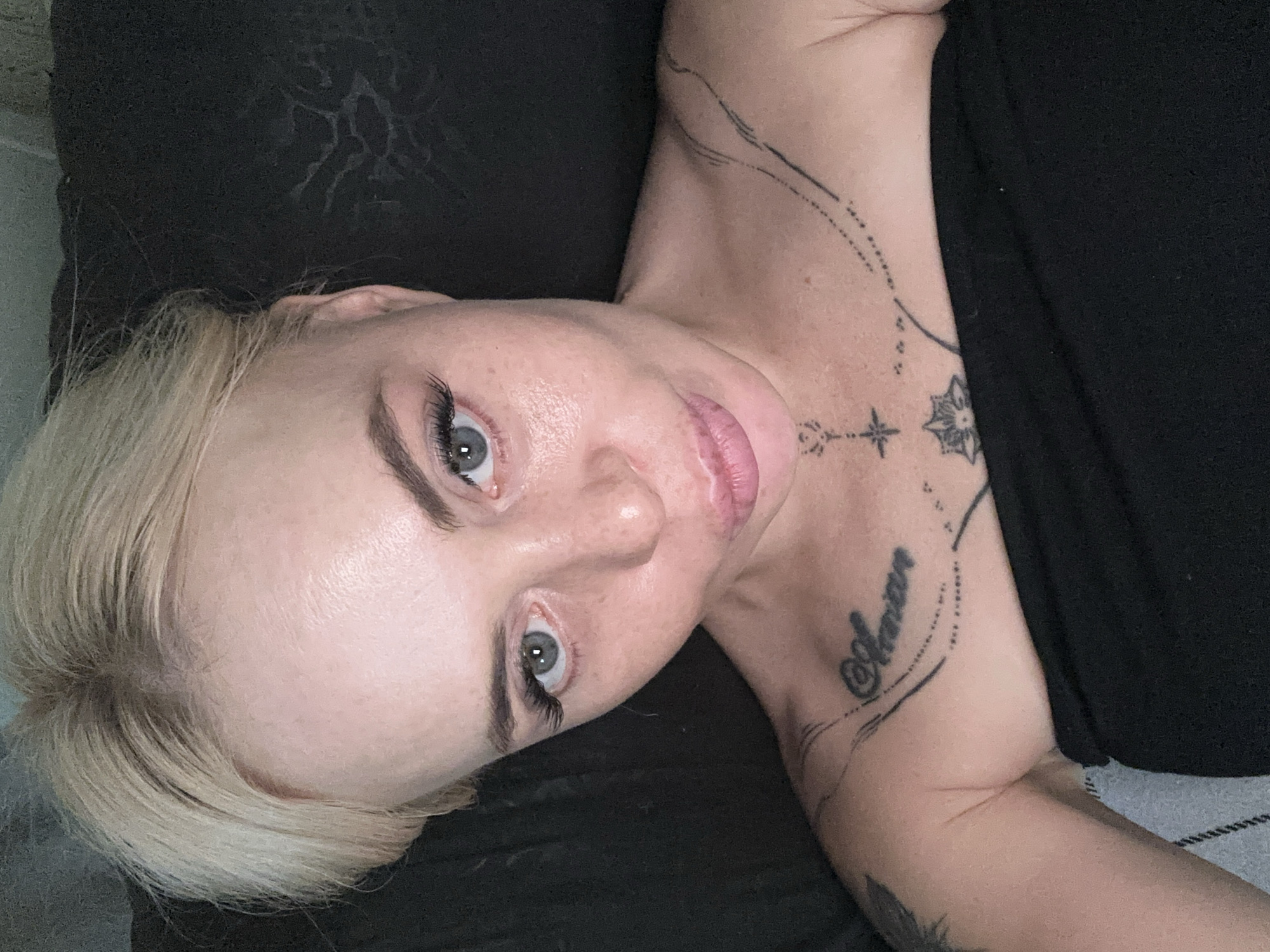 The image is a close-up, high-angle selfie of a person with blonde hair and light skin, looking directly at the camera. The person is wearing a black strapless top that covers their chest. There are visible tattoos on the person's chest and collarbone. The background appears to be a dark, textured surface, possibly upholstery. The lighting is dim, with the face being the primary illuminated area. The person's facial expression is a slight pout, with eyes looking forward and eyelashes appearing to be enhanced. There are no identifiable environmental features to place this scene in a specific city or location.