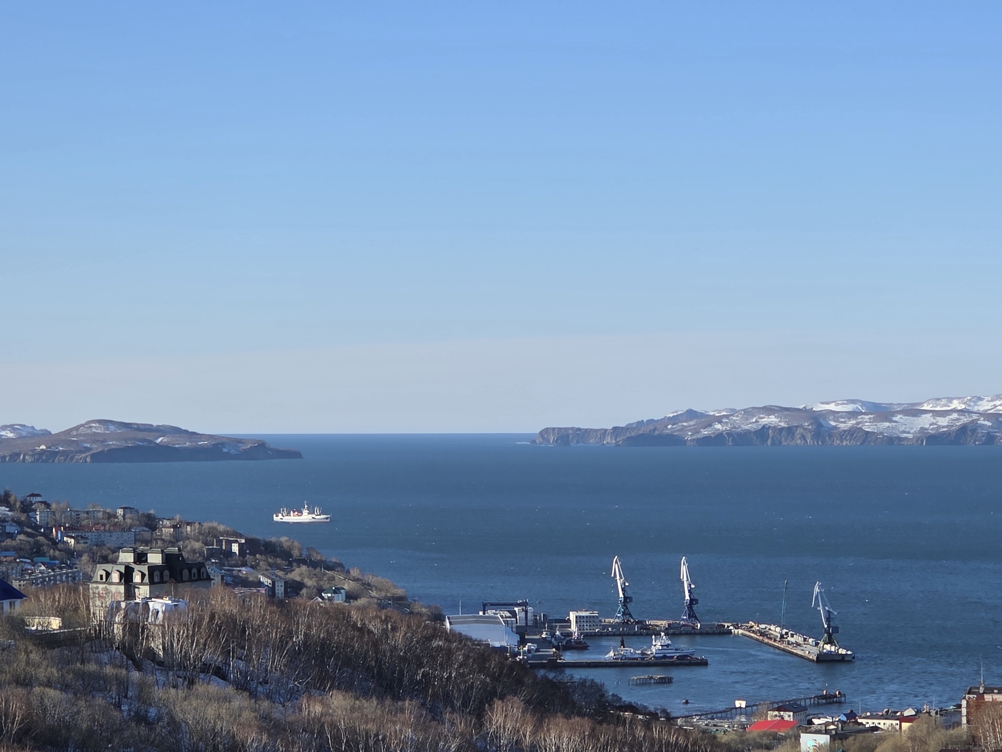 The image depicts a cityscape and harbor on a clear day in an unknown location. In the foreground, buildings cluster on a hillside interspersed with leafless trees, some with visible snow patches. Beyond the city, a harbor features several cranes, docks, and various watercraft. A larger ship is seen at sea in the midground. In the distance, islands rise from the water, some with visible snow-covered peaks. The sky is a clear, pale blue.