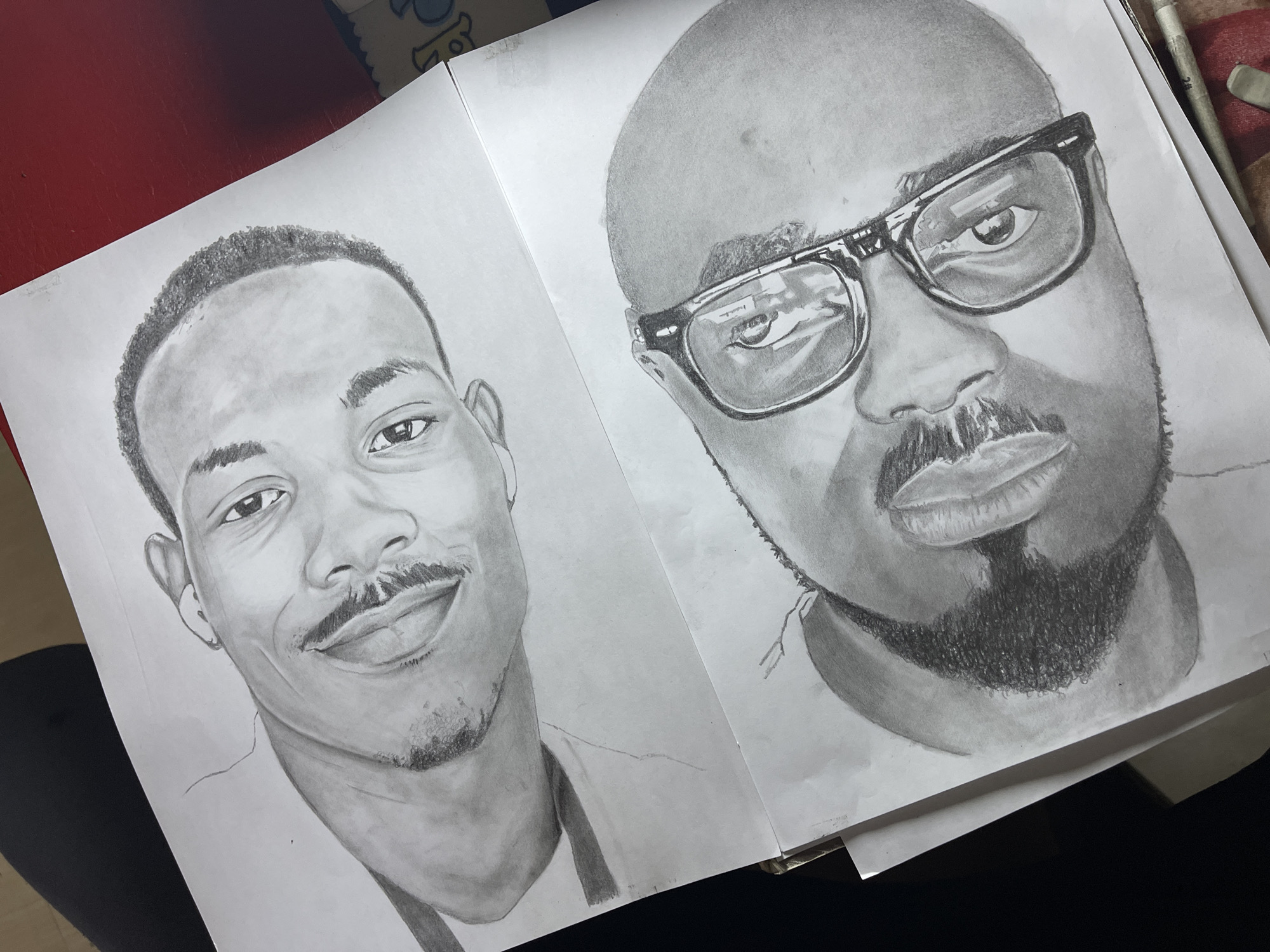 Two pencil portraits are displayed side-by-side on white paper, likely a sketchbook or art pad. The portrait on the left depicts a man with a dark complexion, a slight mustache, and short hair. He is smiling. The portrait on the right shows another man, also dark-skinned, with a shaved head and a thick beard. He is wearing glasses with a distinct reflection in the lenses. The environment is indeterminate, appearing to be a tabletop or workspace. Jos, Nigeria is not discernable from the image.