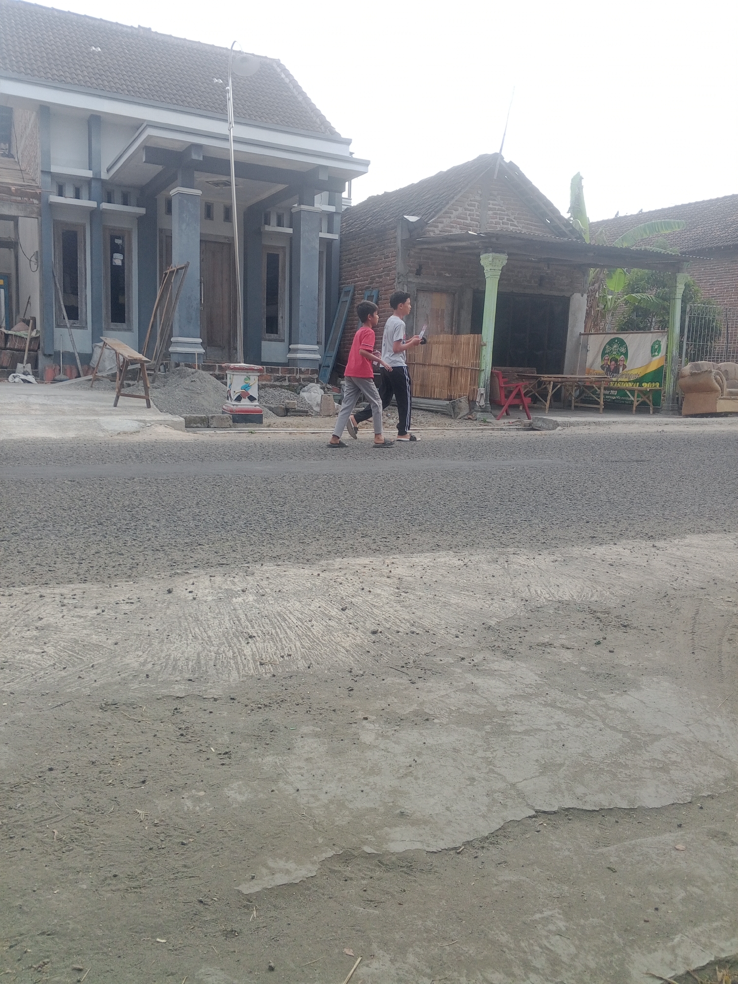 The image depicts a street scene in Lembah, Indonesia. Two young boys are walking on the right side of the street. The boy on the left is wearing a red shirt and grey pants, while the boy on the right is wearing a white t-shirt and black pants. They are walking away from the viewer and towards the right side of the frame. In the background, there are two houses and some construction materials. The house on the left appears to be under renovation, with scaffolding and piles of sand visible. The house on the right has a more rustic appearance with a corrugated metal roof. A banner with text and images is hanging on the front of the right house. The sky is overcast. The street is paved with asphalt, and there is a concrete area on the left side of the street which shows signs of wear and tear, with cracks and uneven surfaces.
