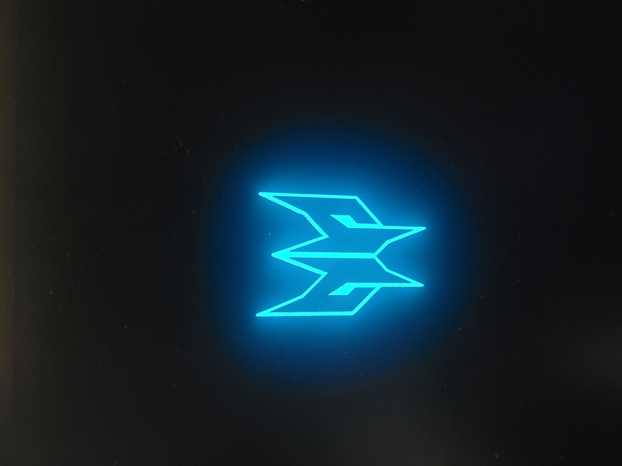 A blue neon logo is displayed against a dark background. The logo is symmetrical and appears to be made of sharp, angular lines, resembling a stylized wing or a bird of prey. The neon light creates a bright glow around the logo, with a soft blue halo emanating outwards. The background is a deep, dark color, possibly black or a very dark grey, which allows the illuminated logo to stand out prominently. The surface appears matte, with some subtle texture or specks visible upon close inspection.