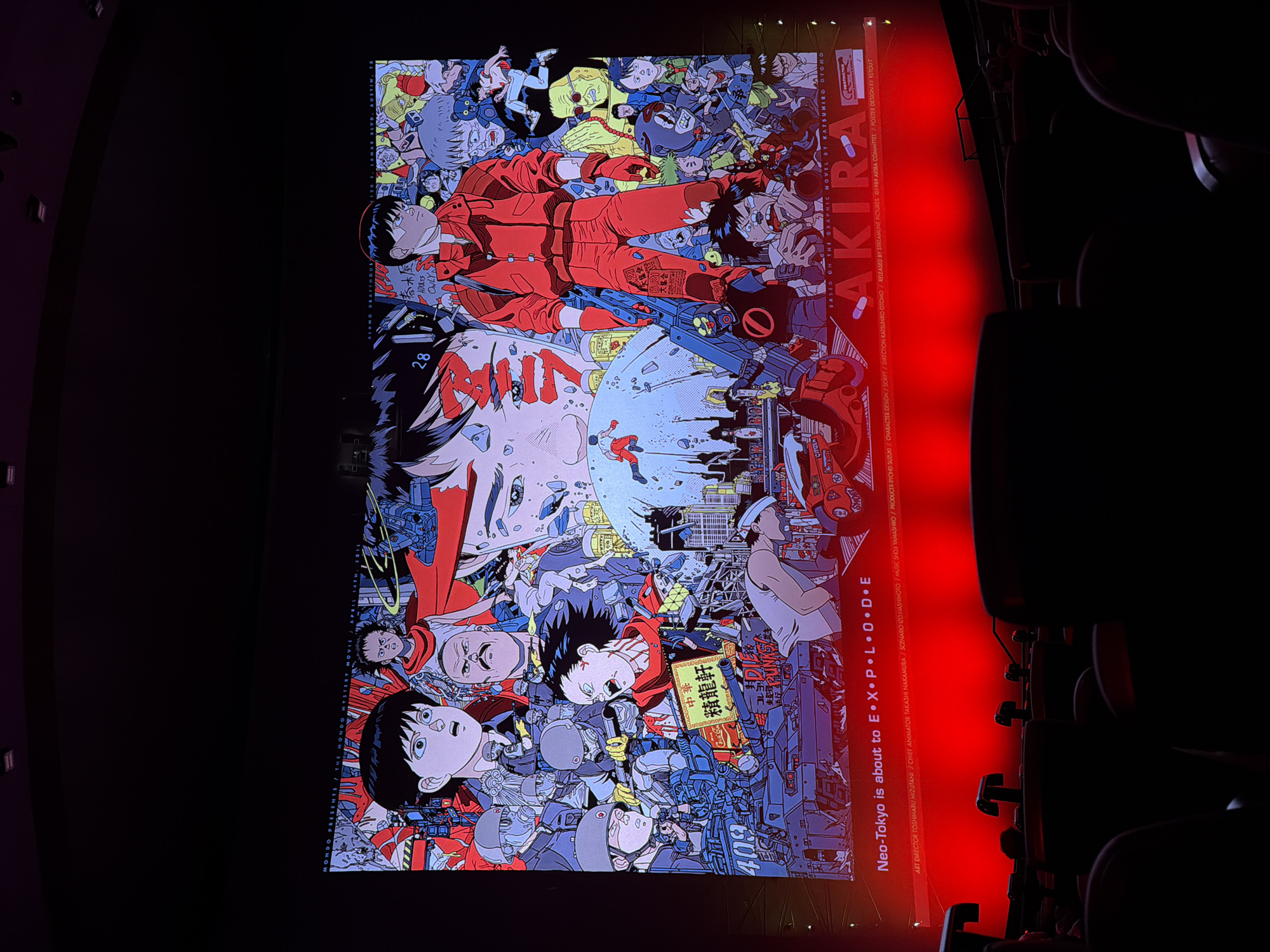 A photo taken inside a dark movie theater shows a screen displaying a poster for the animated movie "Akira." The poster features a collage of characters and scenes from the film in a chaotic composition dominated by blues, reds, and whites. A red strip on the right side of the screen contains white text listing the movie's crew and a tagline: "Neo-Tokyo is about to EXPLODE." Rows of black movie theater seats are visible on the right side of the image, indicating the photograph's setting.