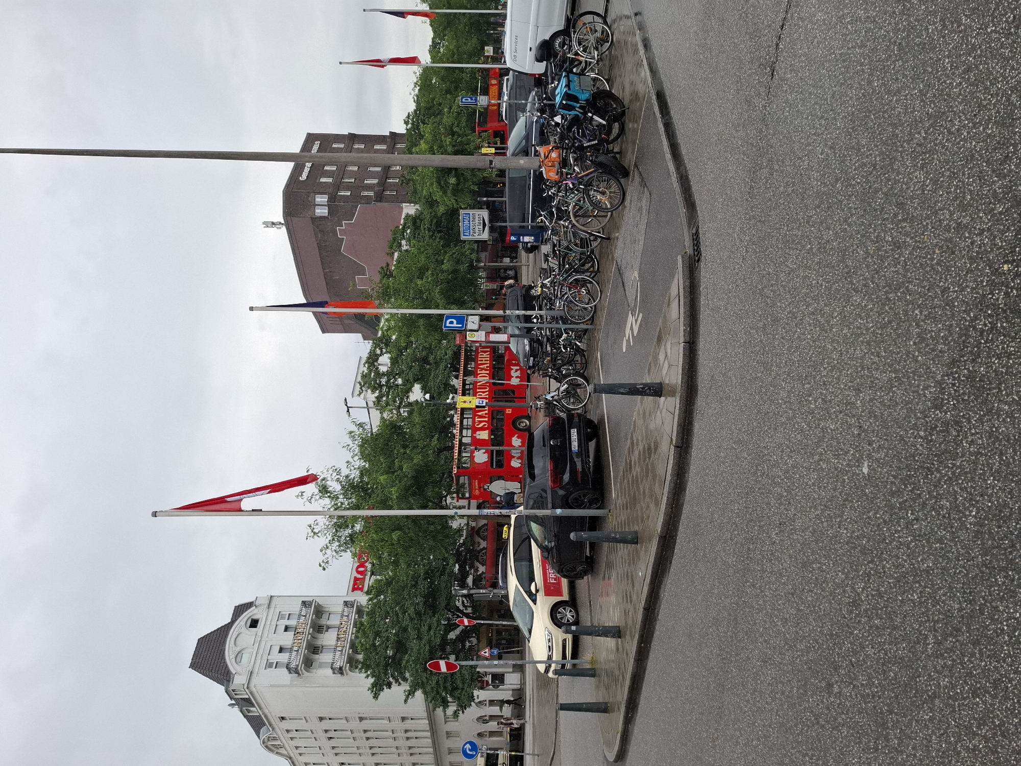 The image depicts a street scene in Hamburg, Germany, likely on a cloudy or rainy day as indicated by the wet asphalt and overcast sky. In the foreground, a paved road curves to the left, with a raised sidewalk or curb area. Several bollards are placed along the edge of the street.
On the sidewalk area, a black car, identified by license plate "HH DU 1964", is parked. Next to it, a white taxi with "FREI" written on its door is also parked. A considerable number of bicycles are parked in a designated area behind the cars, some with saddlebags or baskets.
In the background, a red double-decker bus, labeled as a "STADTRUNDFAHRT" (city tour), is stopped. Adjacent to the bus is a multi-story building with a brick facade and multiple windows. To the left of the bus, a white, multi-story building with a distinctive mansard roof and ornate balconies is visible. Several flags are flying from flagpoles in the scene, including what appear to be German and possibly other European flags. Signage indicates parking areas, with one sign reading "AUTOMAT PARKSCHEIN hier lösen" (Pay for parking ticket here). Trees line the street, contributing to the urban landscape. There are a few indistinct figures of people visible walking in the background. A white utility van is parked further down the street.