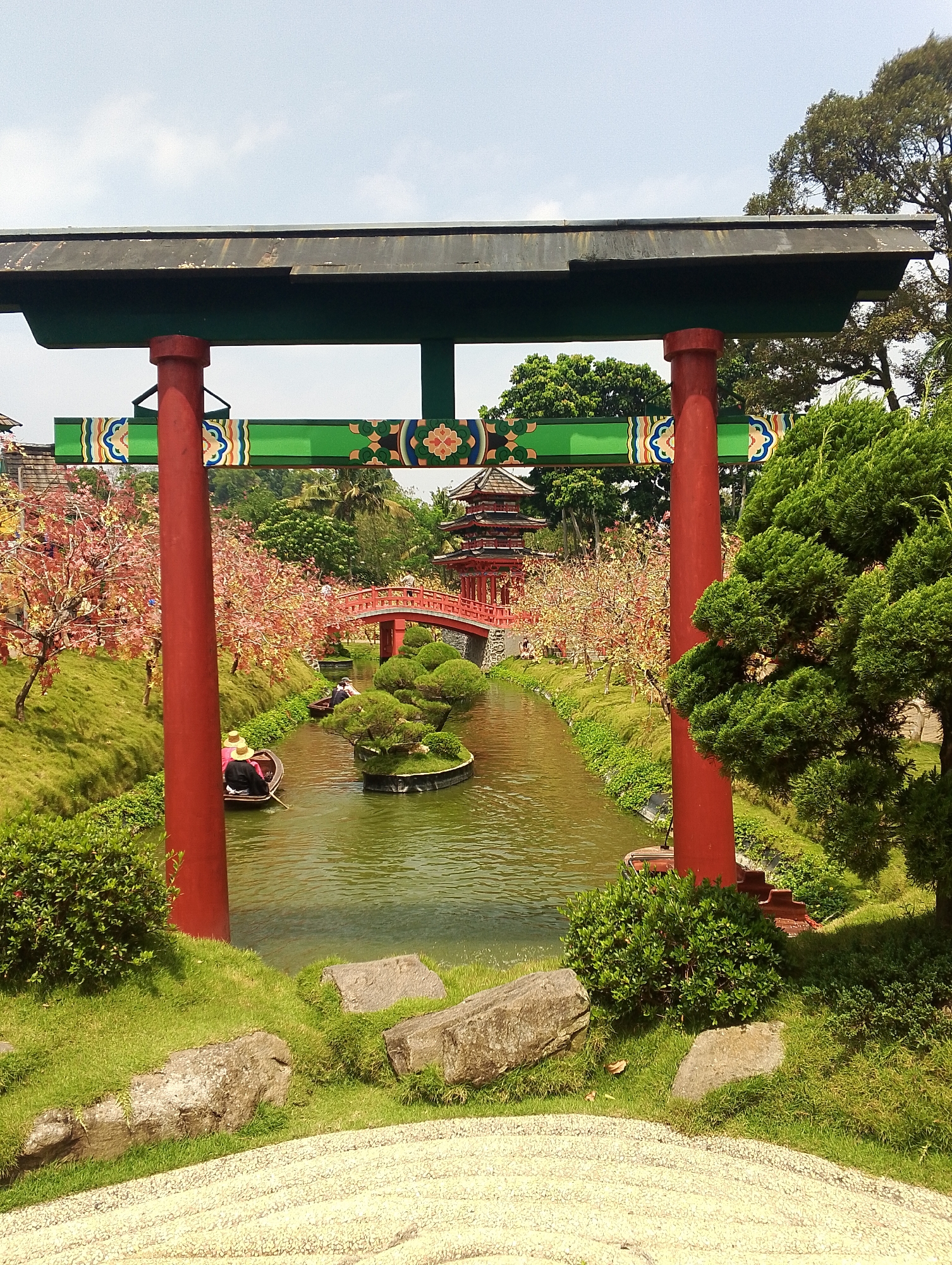 The image depicts a scenic view of a Japanese-style garden, likely located in the city of Megamendung, Indonesia. In the foreground, a large, traditional red torii gate stands prominently, framing the scene. Beyond the gate, a tranquil canal winds through the garden, its water reflecting the surrounding greenery.
Two small boats navigate the canal. In the closer boat, two individuals wearing straw hats are engaged in rowing. Further down the canal, another boat with two occupants is also in motion. The banks of the canal are lush with vibrant green grass and meticulously sculpted bushes, some resembling small islands.
On the left side of the canal, trees with delicate pink blossoms add a touch of floral beauty to the landscape. In the background, a multi-tiered pagoda-style structure and a vibrant red bridge create a distinctly East Asian architectural aesthetic. The entire scene is bathed in bright daylight, with a clear sky overhead, indicating fair weather. The presence of sandy, raked ground in the lower foreground suggests a deliberate Japanese garden design element.