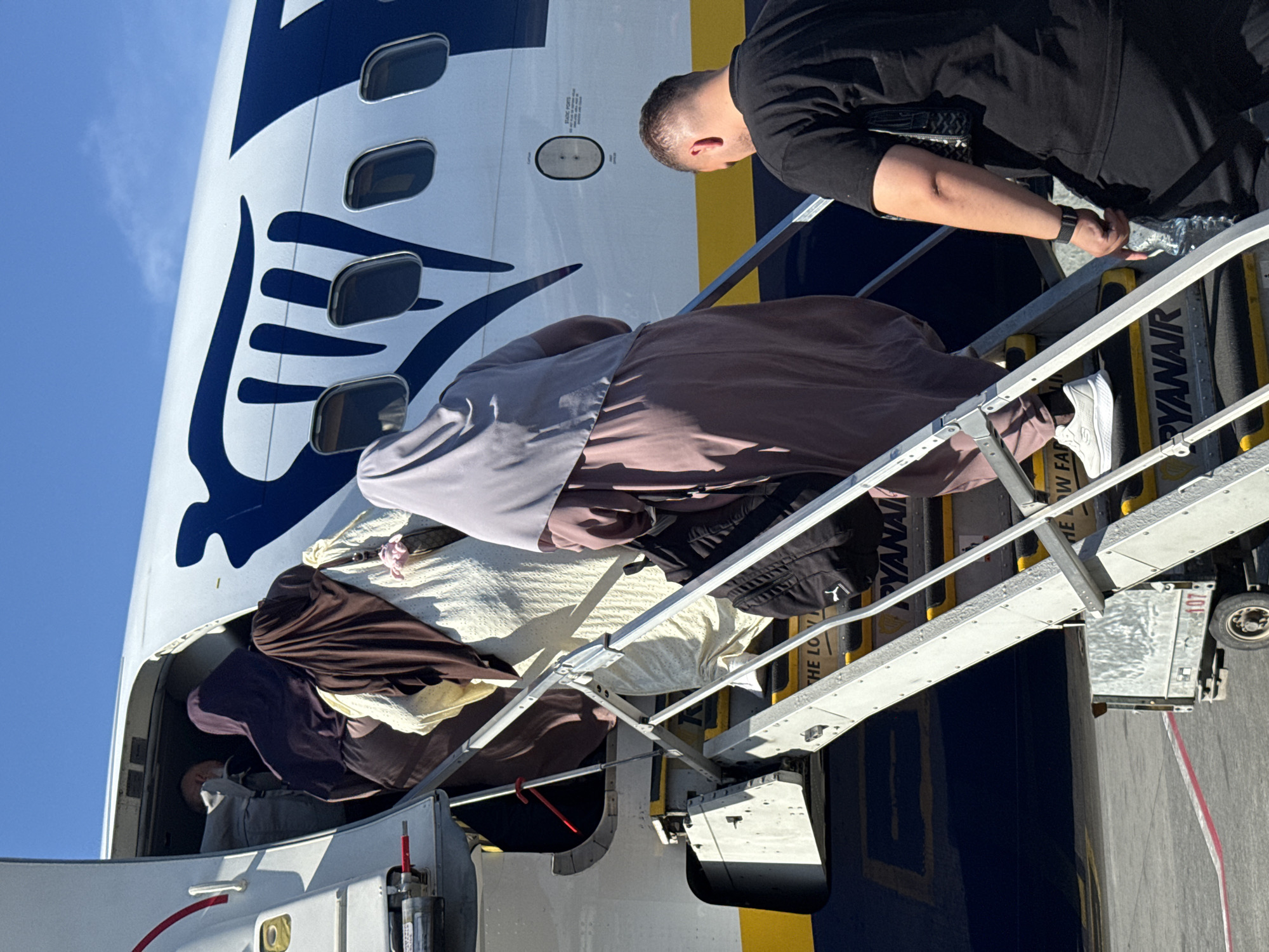 In the city of Kruja, Albania, on a sunny day, a group of people are boarding a Ryanair airplane via a portable staircase. The plane's fuselage is white with a large dark blue graphic. Several individuals, predominantly women wearing hijabs in various colors including purple, brown, and grey, are ascending the stairs. One woman in a beige dress is also visible. A man in a black t-shirt and black pants is descending the stairs, holding a bottle of water. The staircase is made of metal with yellow safety markings on the steps, and the Ryanair logo is visible on the steps. A small utility cart with the number 107 is positioned at the bottom of the stairs. The ground appears to be tarmac.