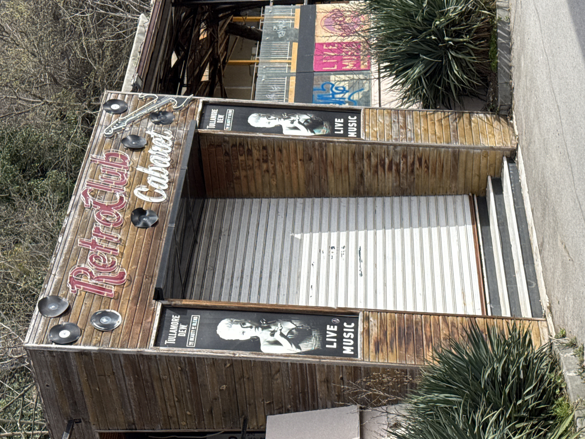 A still, slightly off-kilter, and high-angle shot depicts the exterior of the "Retro Club Cabaret" in Varna, Bulgaria. The building is constructed of wood. The facade features the name of the club in vintage lettering, adorned with record-shaped decorations and a guitar-shaped ornament. The entrance is secured with a rolled-down white metal shutter. Banners advertising "Tullamore Dew" with an image of a female figure and the phrase "Live Music" are displayed on either side of the entrance. Above the structure, partially visible, are metal beams. To the side of the building are green plants. A concrete sidewalk is seen on the right. In the background are trees and graffiti.
