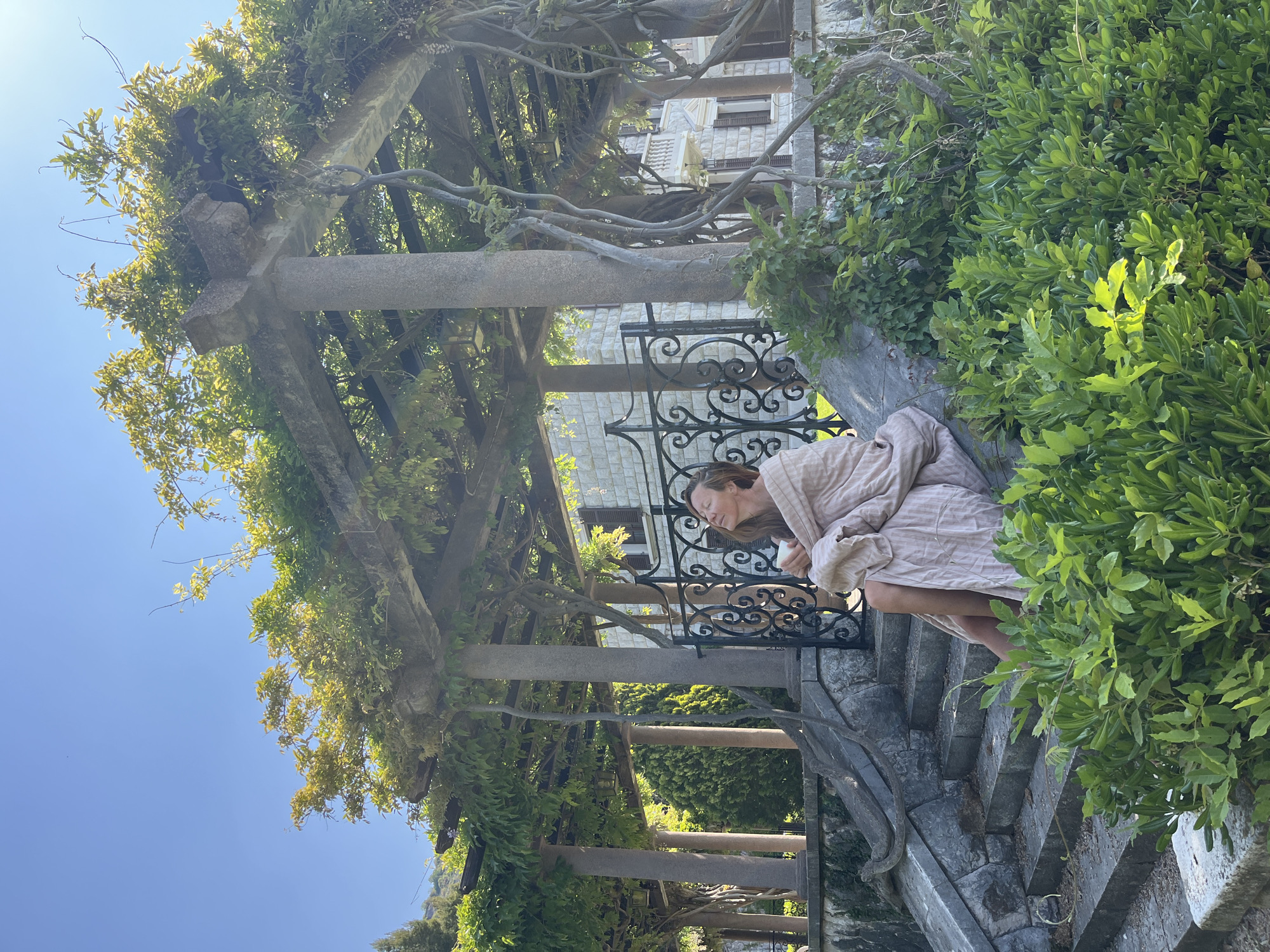 The image captures a serene outdoor scene, likely a terraced garden or a secluded balcony in Sveti Stefan, Montenegro, under a clear, bright blue sky. A woman is seated on rough-hewn stone steps or a low wall on the right side of the frame, facing slightly left and looking downwards with her eyes closed or cast low. She is dressed in a light-colored, possibly striped, loose-fitting garment, resembling a robe or light dress, and holds a small white object, perhaps a cup or phone, in her hands, suggesting a moment of contemplation or relaxation.
The scene is dominated by an intricate stone pergola or archway structure with thick, sturdy pillars and beams, heavily overgrown with various lush green vines and foliage, creating a natural canopy. An ornate black wrought-iron railing is visible behind the woman, separating her from a light-colored building in the mid-background. This building features stone or tiled walls and multiple windows with dark frames, topped by a distinctive, intricate roof structure. The ground around the woman and the structure consists of rugged stone steps, also partially covered in greenery. The bright sunlight casts distinct shadows, indicating a clear, sunny day. No text is visible in the image.