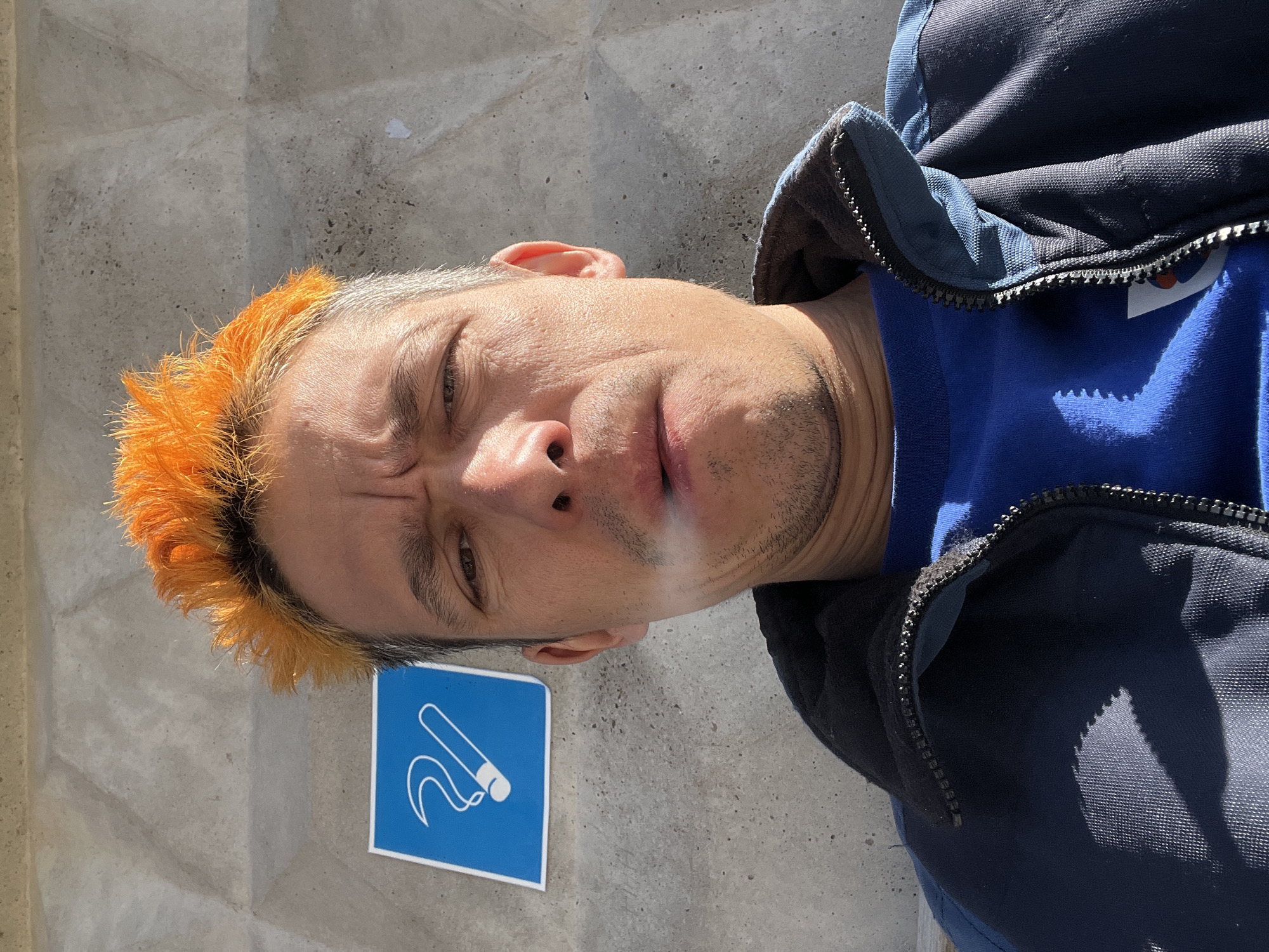 The image depicts a man with orange hair smoking a cigarette, possibly standing in a designated smoking area. A blue sign with a white cigarette graphic indicates this possibility. The man is wearing a blue shirt and a dark blue jacket. The background is a textured concrete wall. The picture appears to have been taken outdoors in Chaikovsky, Russia.