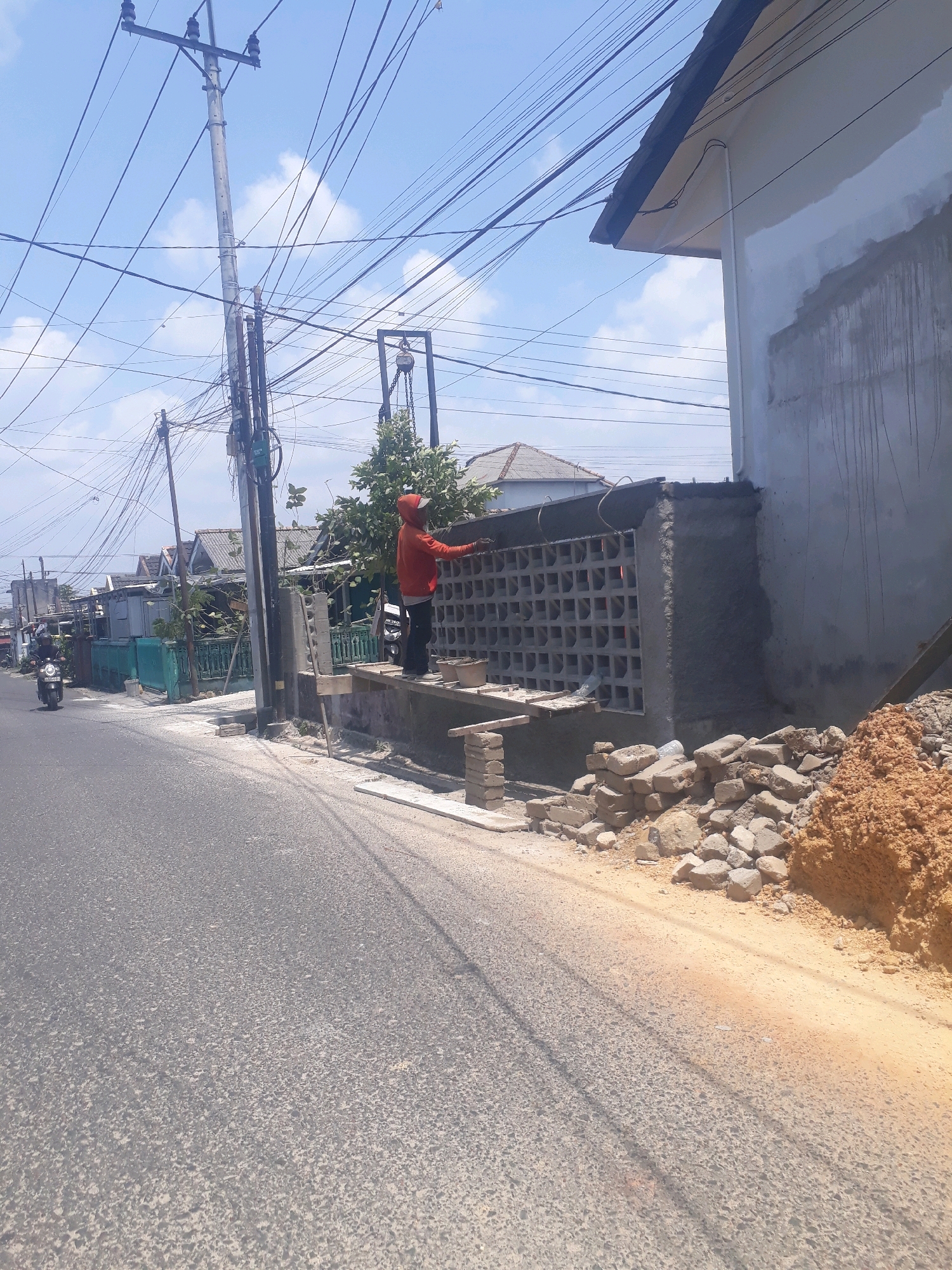 The image depicts a street scene in Pangkalpinang, Indonesia, under a clear sky with scattered clouds. A construction worker, wearing an orange hooded jacket and black pants, is actively engaged in building a wall. The wall features a decorative pattern of square openings and is in the process of being cemented. The worker is standing on a temporary wooden platform, and various construction materials like bricks, cement bags, and a pile of soil are visible nearby. To the left of the construction site, a paved road runs parallel to the sidewalk. A motorcycle carrying a rider is seen on the road, heading away from the camera. In the background, residential houses and electrical poles with numerous wires are scattered across the landscape, indicating a typical urban or suburban environment. The overall scene suggests ongoing construction work on a sunny day in Pangkalpinang.
