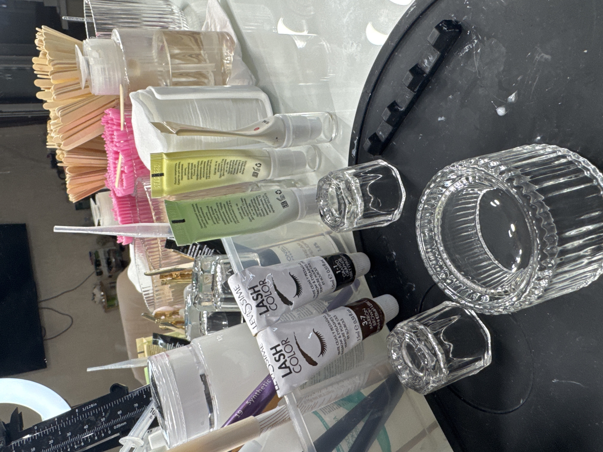 The image depicts a close-up, top-down view of a tabletop cluttered with various beauty and cosmetic products. The scene appears to be set indoors, likely in a personal workspace or a salon, given the array of items. In the foreground, two clear glass receptacles, one cylindrical and ribbed, the other smaller and octagonal, are prominently placed on a dark, matte surface. Scattered white specks, possibly residue from cosmetic products, are visible on this surface.
The middle ground is filled with an assortment of tubes and containers. Two small squeeze tubes labeled "LEVISSIME LASH COLOR" are visible, one in brown and the other in graphite, suggesting products for tinting eyelashes and eyebrows. Alongside these are other tubes with colorful labels, one light green and another yellow, both bearing Cyrillic text, indicating they are likely cosmetic or personal care items. A clear plastic bottle with a pump dispenser, containing a light-colored liquid, is also present. A white, folded item, possibly cotton pads or tissues, sits in a white holder. A thin, metallic applicator with a pointed tip is also visible.
Further in the background, a large number of wooden sticks, resembling spatulas or applicators, are arranged in a fan-like cluster. Several pink bristled brushes are also visible. A clear, ribbed container holds a transparent liquid. The background is somewhat blurred, but it suggests a reflective surface, possibly a mirror or a glass partition, reflecting a ring light and other indistinct objects. A black ruler with white markings is partially visible on the left side of the image, with its scale indicating measurements up to 70mm.
The image does not provide any direct information about a specific location in Saratov, Russia, or any identifiable individuals. The scene focuses solely on the arrangement of objects on a tabletop.