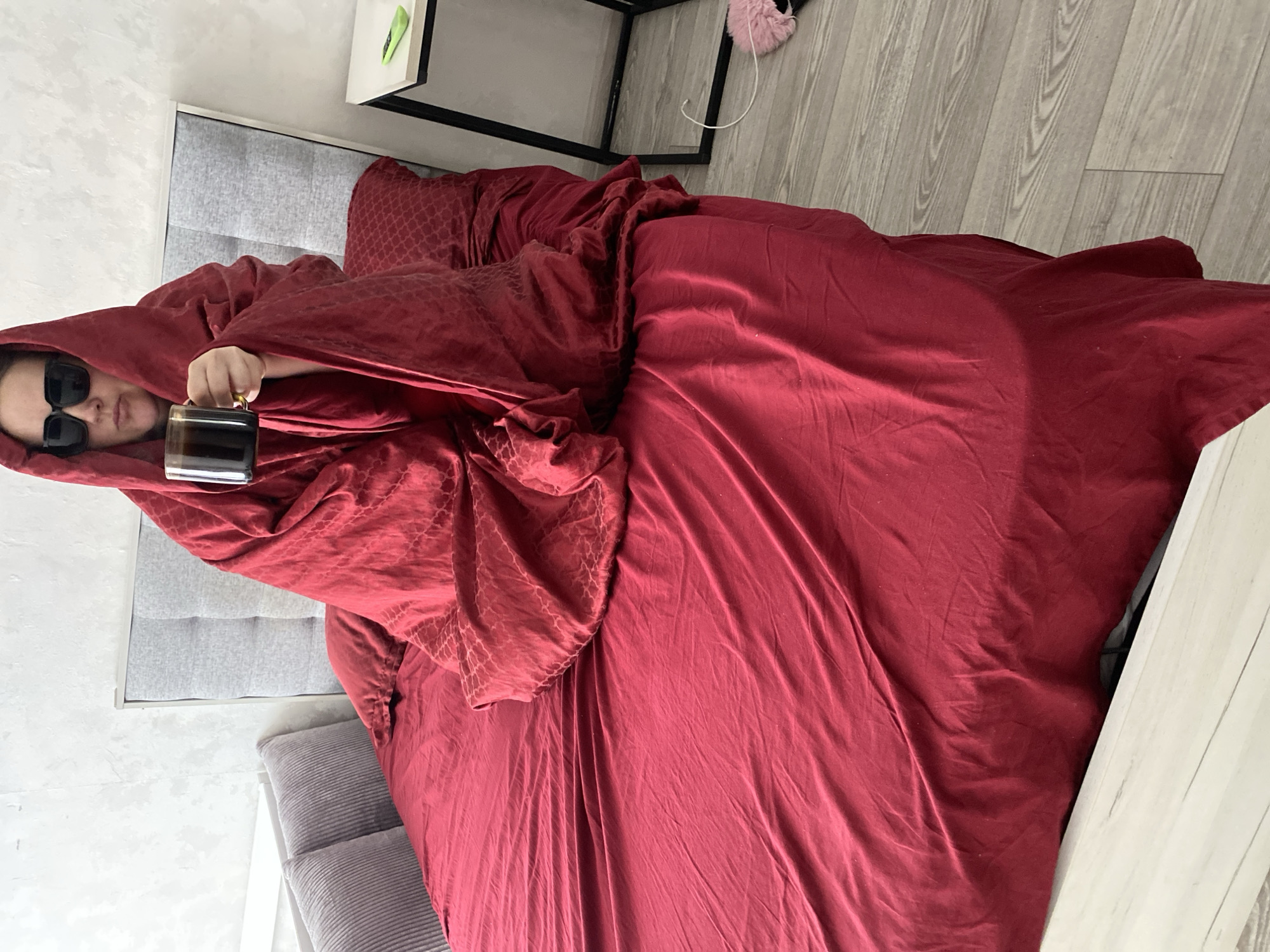 A person wearing sunglasses is lying in a bed covered in a red blanket and holding a mug. The bed has a grey headboard. A small table with a white drawer is next to the bed. The room has a light grey floor. The city of Sochi, Russia is not discernible from the image.