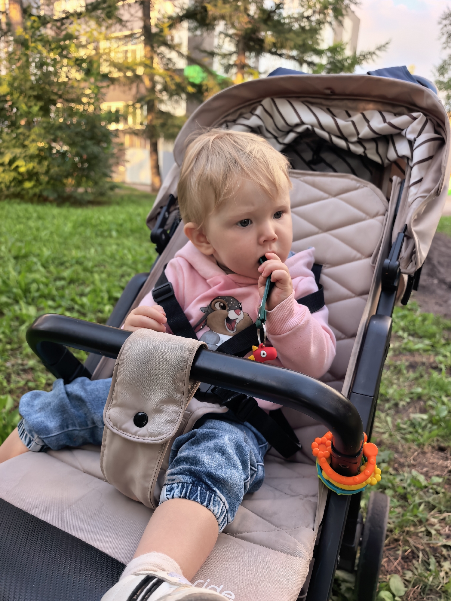 A blonde-haired toddler wearing a pink hoodie with a cartoon squirrel graphic and denim jeans sits in a beige stroller. The child is holding a green toy in their mouth, with a red animal-shaped toy attached to it. A colorful teething toy is attached to the stroller's handle. The stroller is positioned on a grassy area, with trees and a building visible in the background. The scene appears to be outdoors in a park or residential area, possibly in the city of Sterlitamak, Russia, given the time of day suggested by the warm, golden lighting and the presence of mature trees.