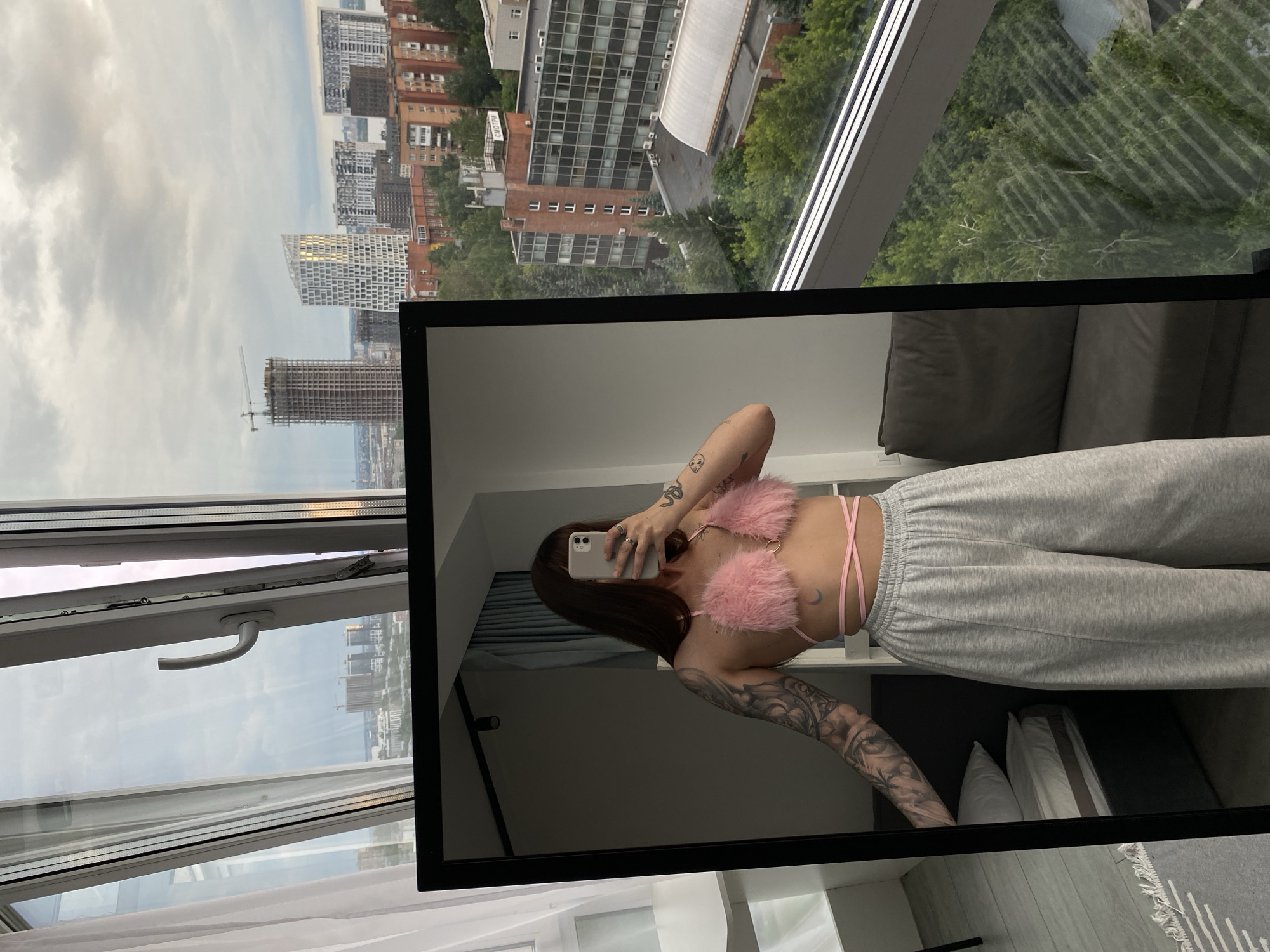 A woman is taking a selfie in a mirror, her reflection showing her torso wearing a pink fuzzy bralette and gray sweatpants. She has tattoos on her arms and a crescent moon tattoo on her back. The setting appears to be an apartment with a large window overlooking a city skyline. The view through the window shows numerous buildings, some under construction, and trees. The time of day is indeterminate, but the lighting suggests it could be daytime. The weather is overcast with scattered clouds. The text "СМОТР" is visible on one of the buildings in the background. The overall mood is casual and self-focused.