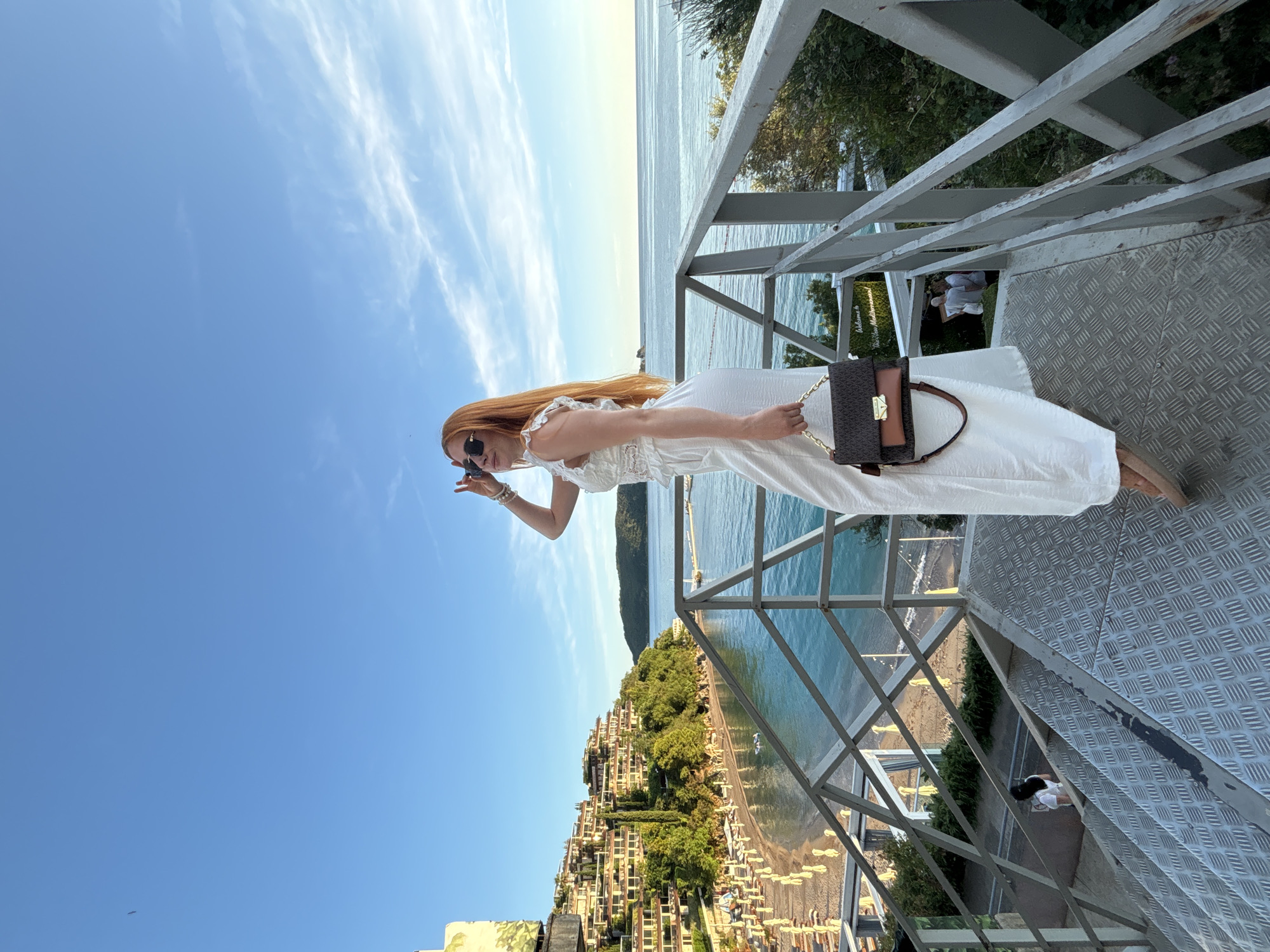 The image captures a vibrant coastal scene from a high vantage point, with the perspective rotated 90 degrees clockwise. The main subject is a woman with long, reddish-blonde hair, dressed in a white, off-the-shoulder maxi dress. She wears dark sunglasses and holds a brown purse with a gold chain strap. Her left hand is raised towards her face, possibly adjusting her sunglasses or making a gesture.
She stands on a metallic, diamond-plate textured platform, which is part of a larger metal structure or balcony extending over the coastline. Metal railings are visible around the platform, providing safety and framing the view.
The background reveals a picturesque setting. Below the platform, there's a bustling beach lined with numerous sun loungers and umbrellas. Beyond the beach, the turquoise-blue sea stretches towards the horizon under a clear, bright blue sky with wispy white clouds. On the opposite side of the beach from the viewer, a series of multi-story buildings, characteristic of Mediterranean architecture, cascade down a verdant hillside, interspersed with lush green trees. In the distance, a large mountain range borders the sea.
The scene suggests a sunny daytime, likely late morning or afternoon, given the bright illumination. The overall atmosphere is one of leisure and scenic beauty, typical of a vacation destination like Budva, Montenegro.
Visible text on a sign in the background, partially obscured by a railing, reads "Welcome to" followed by blurred text that appears to be "Your New Mediterranean" or similar wording. Another blurred figure is visible on the beach pathway below the platform.