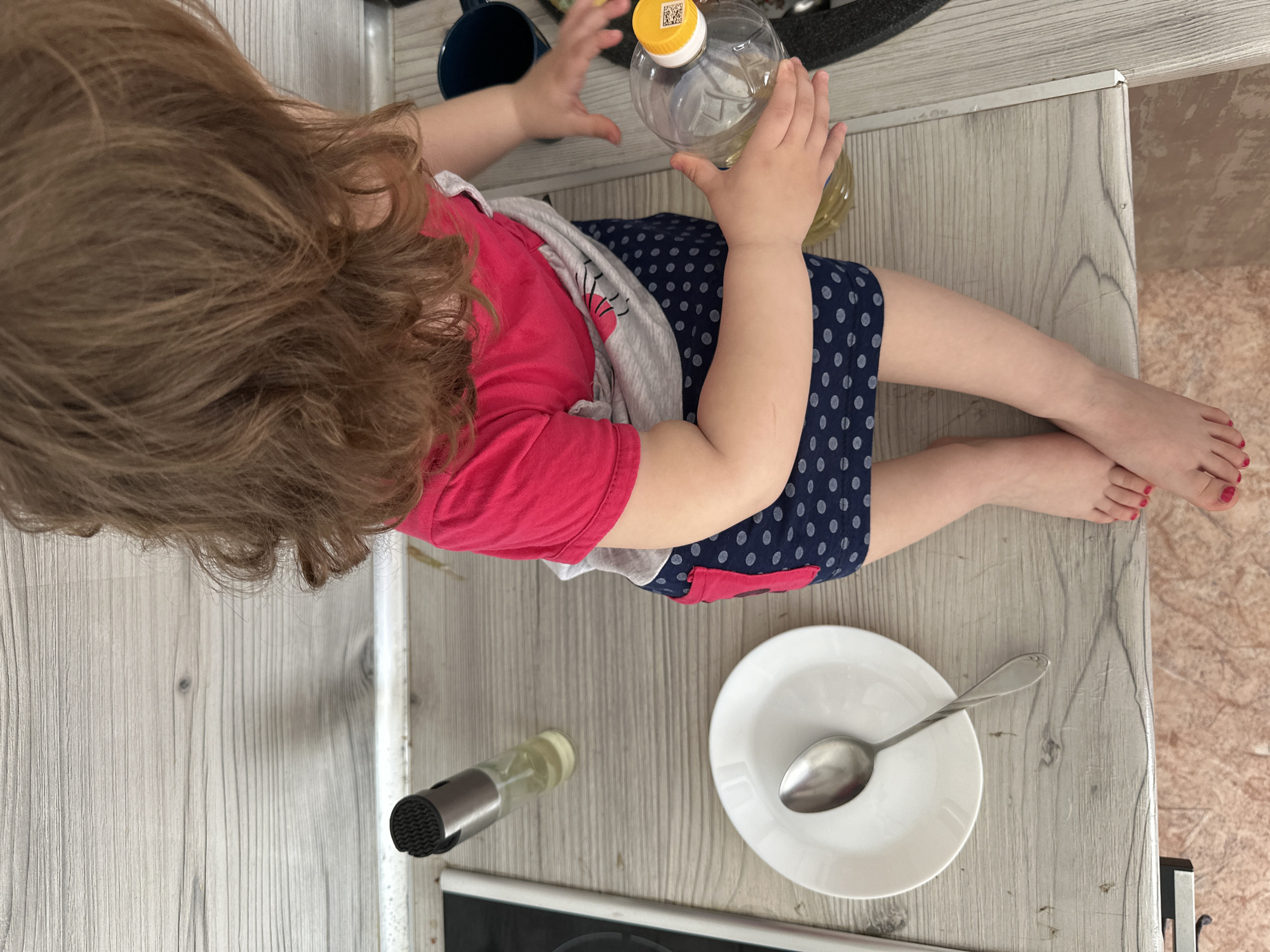 A child with light brown hair is sitting on a kitchen counter in what appears to be a domestic environment, possibly in the city of Chaikovsky, Russia. The child is wearing a pink t-shirt, a grey undershirt, and a blue skirt with white polka dots. The child is holding a plastic bottle with a yellow cap. Near the child are a blue mug, a white plate with a silver spoon, and a glass bottle with a black top. The child's toenails are painted red. The kitchen counter has a light wood grain finish. The floor is a marbled tile pattern.
