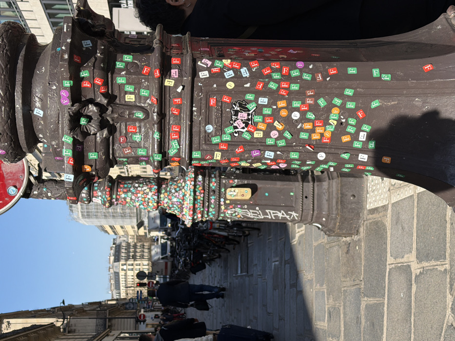 This image depicts an urban street scene in the city of Paris, France, observed during daylight hours. The foreground is dominated by two dark, ornate cast-iron street lamp posts. The closest post on the right is extensively covered with numerous small, square "F" stickers in varied colors, including green, red, orange, purple, white, pink, and yellow; some are peeling. A circular red sign is partially visible at the top of this post. Immediately behind it, a second similar lamp post is also densely adorned with multi-colored "F" stickers, presenting a mosaic-like appearance, and displays some black graffiti and a small rectangular yellow object affixed to its side.
The scene extends down a cobblestone pedestrian pavement, which is partially illuminated by sunlight, creating distinct shadows, and partially in shade. A line of parked bicycles is visible along the left side of the street. Several pedestrians are present on the pavement: a person with dark hair is partially visible on the far right behind the front lamp post; a male figure in a dark jacket and trousers walks away from the viewer further down the street, carrying a dark bag; and another person is partially visible on the left, alongside a dark blue rolling suitcase.
The street is flanked by multi-story, classic Parisian-style buildings with light-colored facades, numerous windows, and intricate architectural details. Scaffolding is visible on some distant structures. Vehicles, including white vans, are present on the street in the background, along with a red traffic light. The sky above is clear and blue.
