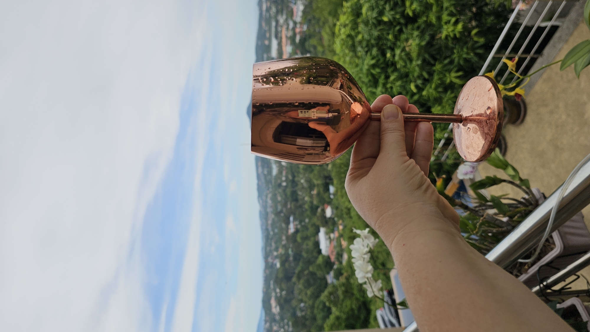 A human hand holds a copper wine glass filled with water droplets against a blurred background of a cityscape. The cityscape appears to be Baan Chaweng, Thailand, with lush green trees, buildings, and a body of water visible in the distance. The sky is partially cloudy with blue patches. In the foreground, a railing and potted plants with white and yellow flowers are also visible. The wine glass itself is reflective, showing a partial reflection of a person holding a phone or camera. The overall scene suggests an outdoor setting, possibly a balcony or terrace with a scenic view.