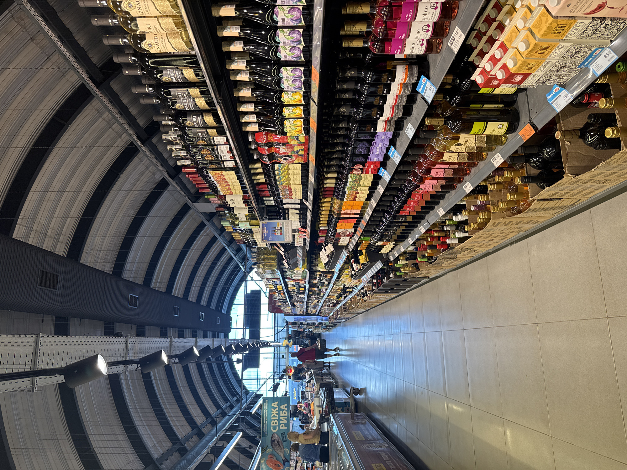This is a wide-angle, eye-level shot of a well-lit, modern grocery store aisle in Dnipro, Ukraine. The aisle runs lengthwise, with shelves stacked high with bottles of wine and other beverages. The store features a distinctive arched ceiling with exposed metal supports and track lighting. On the left side of the aisle, a refrigerated display case runs along the wall, with a sign above it that reads "СВІЖА РИБА" (Fresh Fish). A few shoppers are visible in the distance on the left, browsing or pushing carts.
The right side of the aisle is dominated by towering shelves filled with a wide assortment of wine bottles, organized by color and brand. The bottles are predominantly dark glass, with labels featuring various colors and designs. Below the wine shelves, several cardboard boxes are stacked, containing more bottles, some of which appear to be wine and others possibly juices or other drinks.
In the mid-ground, a man in a red t-shirt walks away from the camera, carrying a basket, facing down the aisle. The floor is tiled in large, light-colored squares, reflecting the overhead lighting. The overall atmosphere is that of a typical supermarket environment, clean and organized, with a focus on alcoholic beverages.