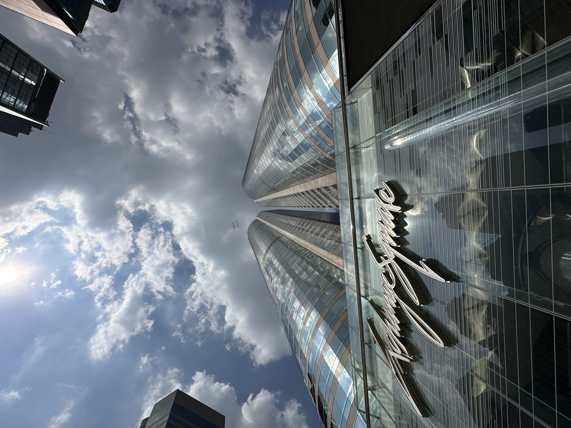 This is a low-angle, wide shot of the "Exchange Square" buildings in Hong Kong. The architecture of the buildings is characterized by their curved glass facades and distinct metallic trim. The sky above is a mixture of blue and dramatic, bright white clouds, with the sun shining brightly and creating reflections on the glass surfaces of the buildings. The "Exchange Square" logo is prominently displayed in a stylized script on the lower glass section of the building in the foreground. The image captures a sense of urban grandeur and modern architectural design.