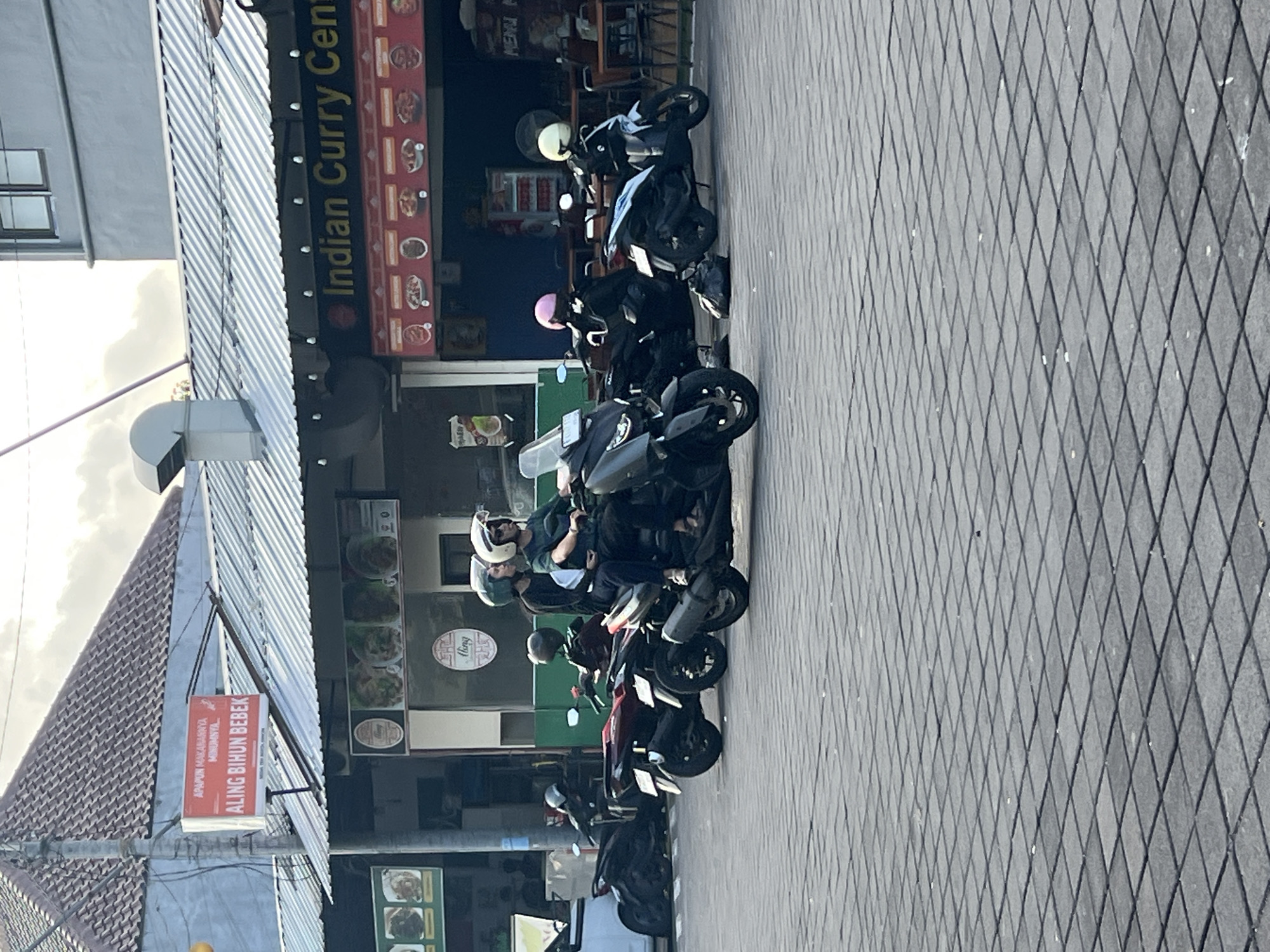 In Kuta, Indonesia, a street scene is depicted with several motorbikes parked and in motion. In the foreground, a grey motorbike with two occupants wearing helmets is the central focus, moving forward. Behind it, other motorbikes are visible, including a red one with a single rider, and several others parked to the left. The background features buildings with awnings, one of which displays a sign in red and white text reading "APAPON MAKANANYA MINUMNYA ALING BIHUN BEBEK". Another awning displays "Indian Curry Cent". The ground is paved with grey interlocking square tiles arranged in a diamond pattern. The overall environment suggests a commercial area in a city.