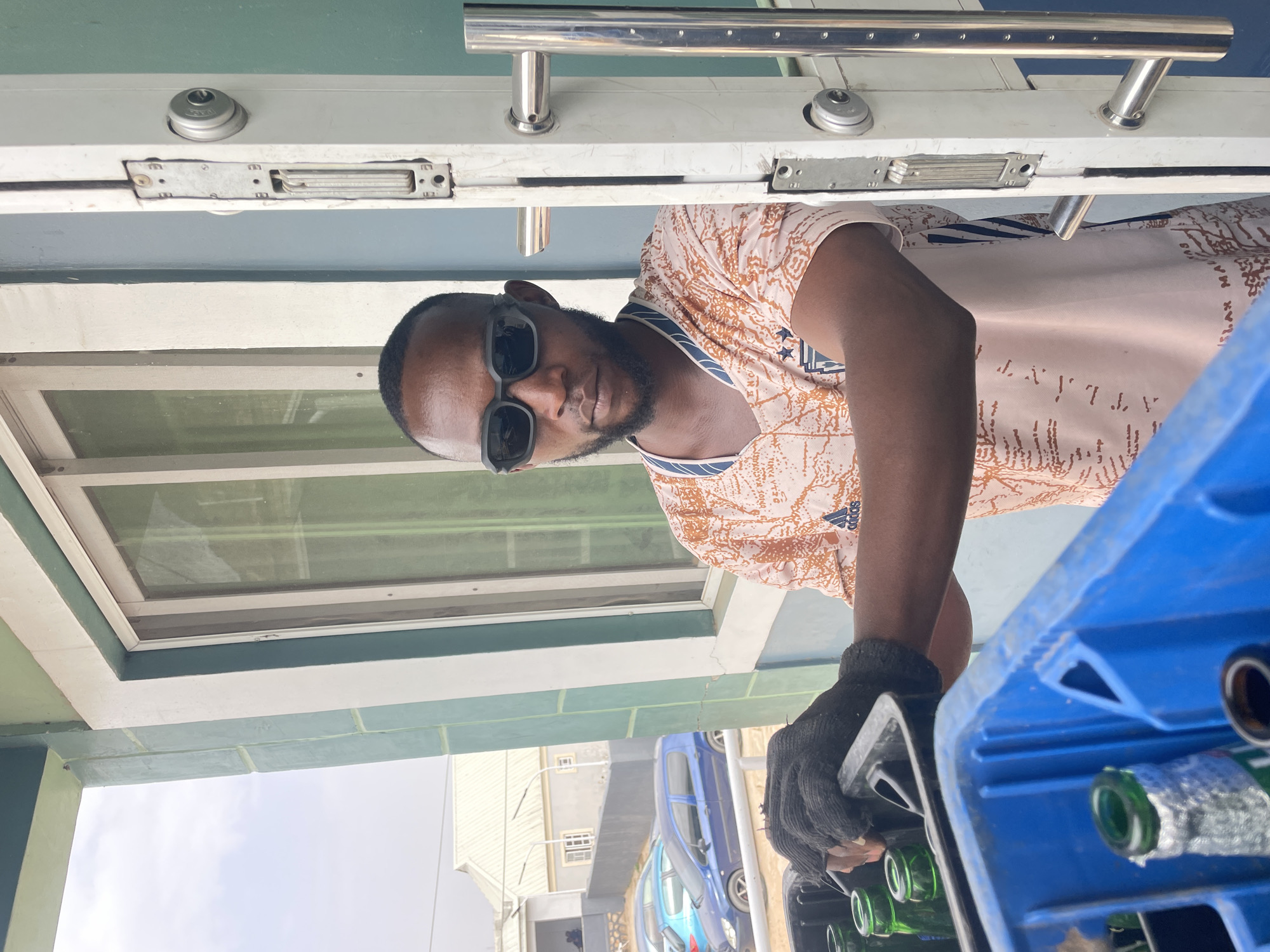 A man with dark skin and a beard wearing sunglasses and a patterned t-shirt is holding a blue plastic crate filled with green glass bottles. He is wearing a grey glove on one hand. He is standing under a structure with a white and green painted exterior, including a window with a screen. In the background are buildings and parked vehicles, including a blue car. The location is Gura Top, Nigeria.