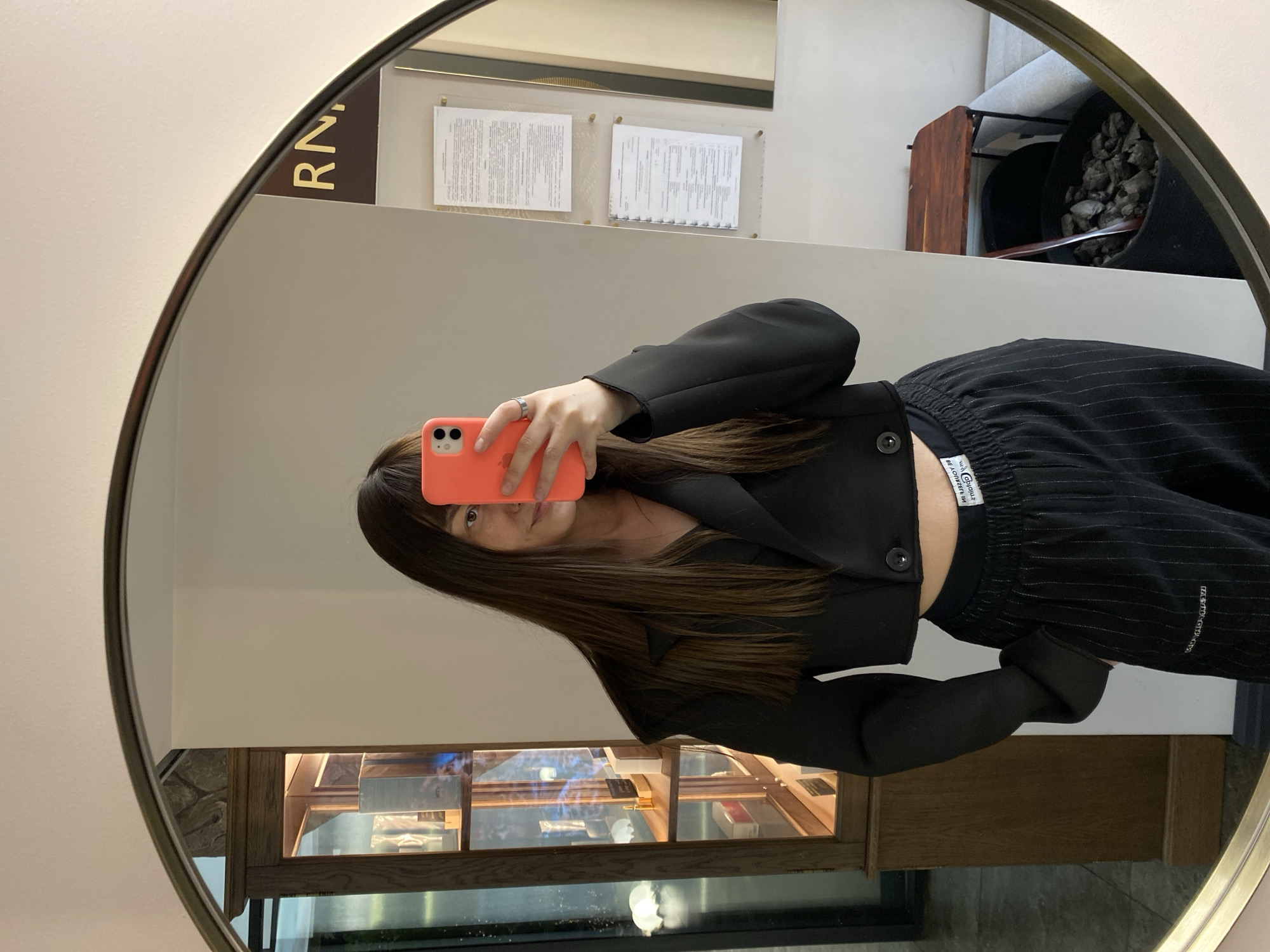 An indoor mirror selfie captures a person holding a coral-colored smartphone, obscuring most of their face except for one eye. The person has long, straight dark brown hair and wears a black cropped blazer/jacket and black pinstriped trousers with an elastic waistband. A silver ring is visible on one of their fingers.
The reflection in the round mirror with a dark frame shows an interior setting. In the upper left, part of a golden sign displays the letters "RN." Below it, two documents are mounted on a light-colored wall in clear acrylic frames; the text on these documents is illegible. To the upper right, a black planter or container filled with dark, rocky material (possibly decorative stones or charcoal) and a wooden scoop is visible.
In the lower part of the reflection, a wooden display case with glass shelves is seen, containing various small items, possibly products or decorations. The overall environment suggests a commercial space like a store, cafe, or reception area, consistent with the provided location context of Votkinsk, Russia.
Notable details on the person's clothing include a black button on the jacket and a white fabric label on the waistband of the pants. Text visible on this label includes "GbciciY" and "WEJOY LIFE" (with "BE JOY" partially visible). The person's stance and the phone's position clearly indicate they are actively taking a mirror selfie.