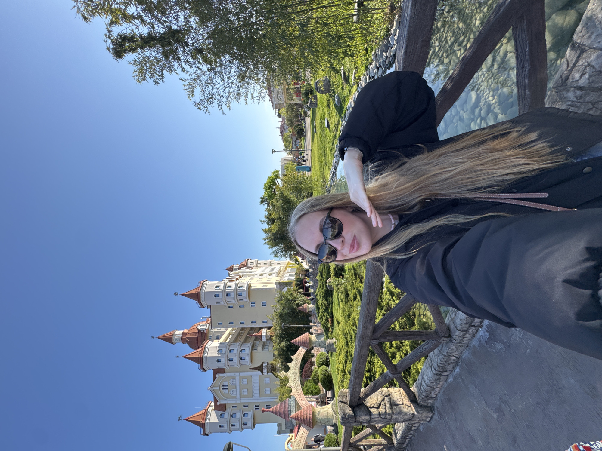 A medium shot depicts a blonde woman wearing sunglasses, standing on a stone-edged bridge with a wooden railing. Behind her is a whimsical, multi-tiered building with orange-roofed towers set against a clear blue sky. Lush greenery surrounds the building, including trees, shrubs, and grass. The location is likely the Sochi Park Hotel in Sochi, Russia, due to the distinctive architecture of the building in the background. The woman appears to be taking a selfie, holding one hand near her face. A small stream runs beneath the bridge.