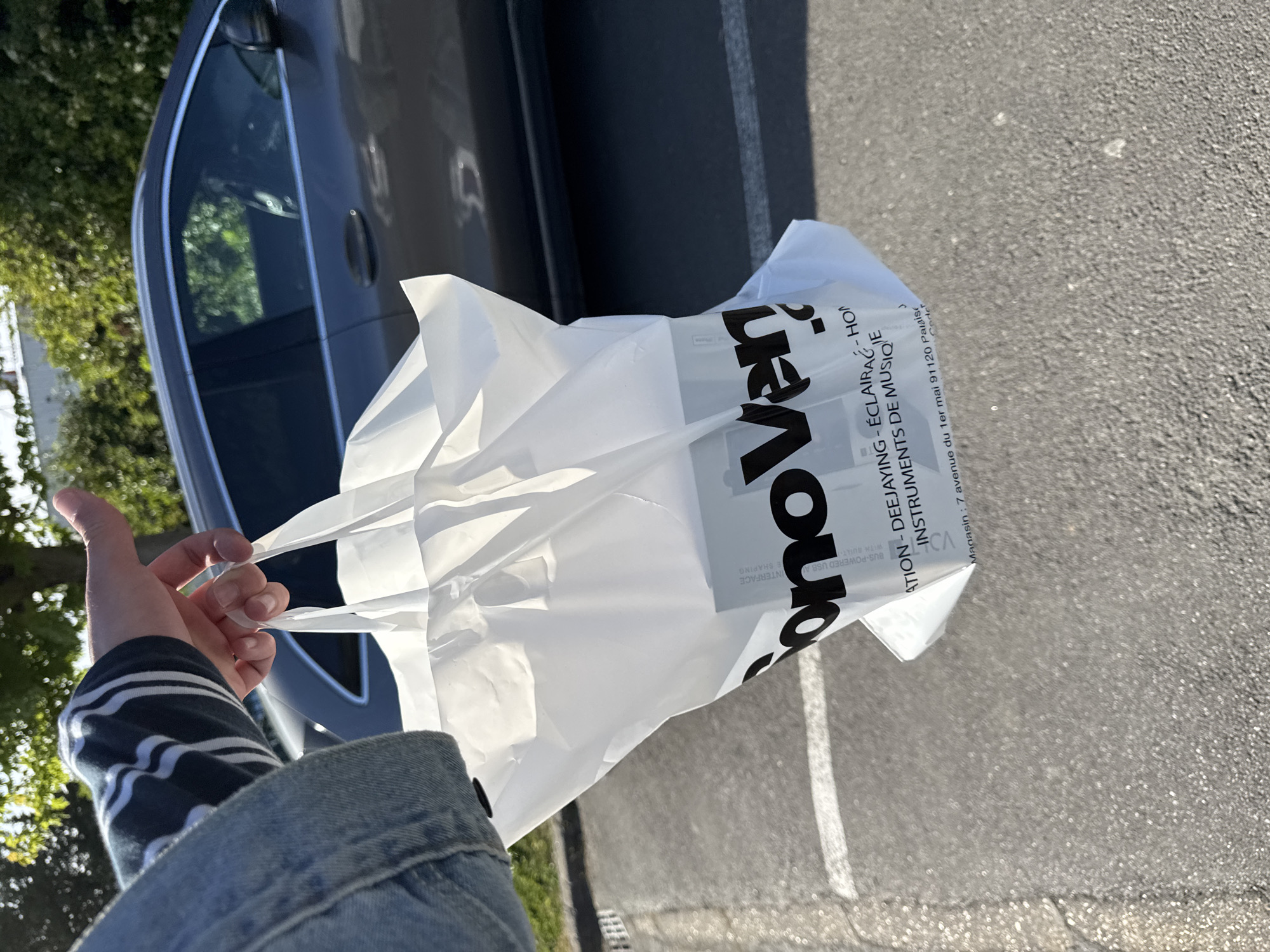 A person holds a white plastic shopping bag from "SonoVente" in their hand. SonoVente specializes in deejaying, lighting, and musical instruments. The bag also has the address "7 avenue du 1er mai 91120 Palaiseau" printed on it. The person is wearing a blue denim jacket over a striped shirt. Part of a gray car is visible behind the bag and the person's hand. The ground appears to be a paved parking lot.