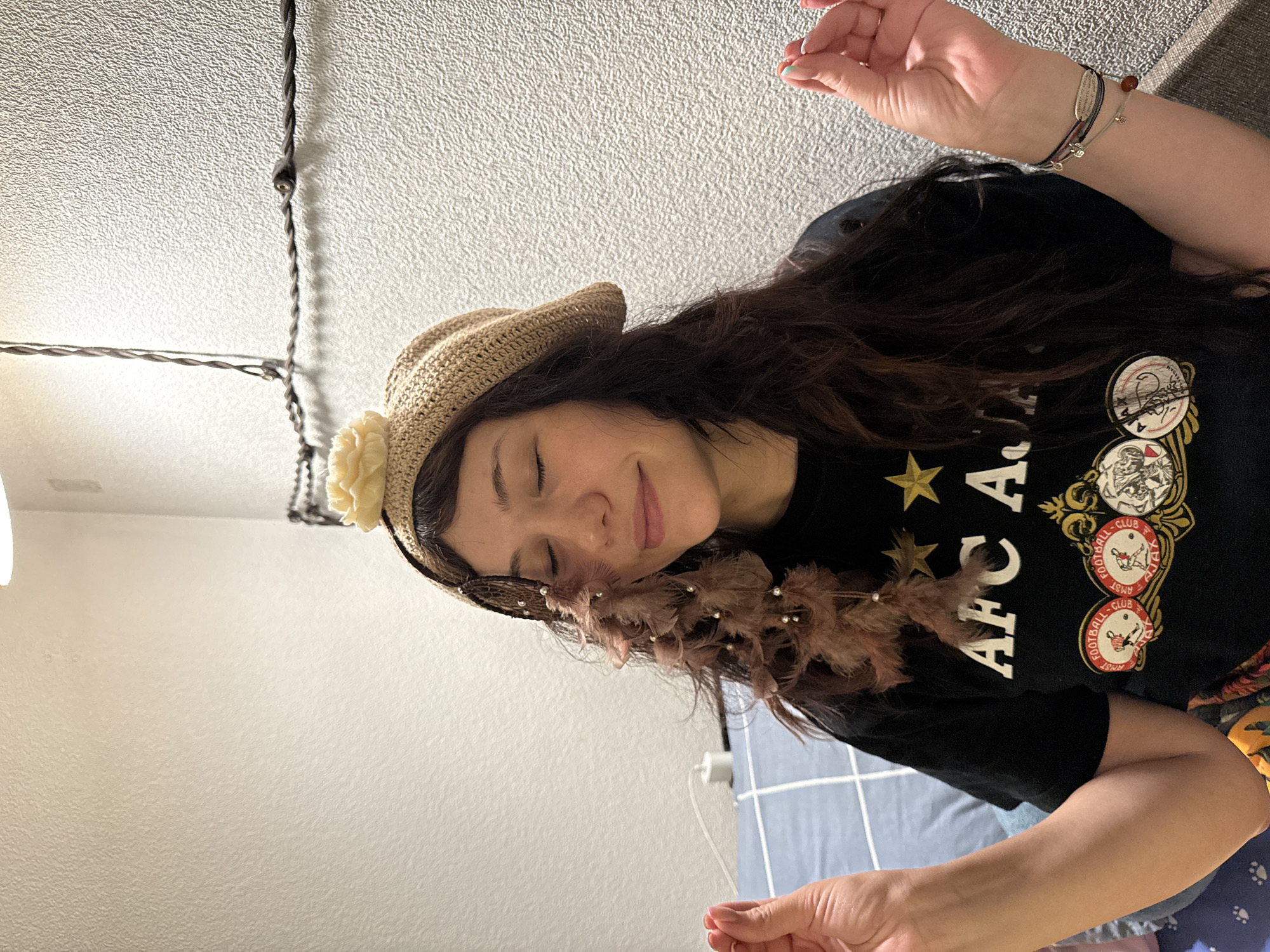A young woman with long, dark, wavy hair is pictured from the chest up, sitting in an indoor setting. She is wearing a light brown crocheted beret adorned with a cream-colored flower and strands of brown feathers and small pearls. Her eyes are closed, and she has a slight smile on her face. She is wearing a black t-shirt with white lettering that reads "AFC ASIA" and features two yellow stars above the text. Below this, there are circular emblems, one of which appears to be a crest with a runner. Her hands are held up with fingers extended, as if in a meditative pose. She is wearing several bracelets on her right wrist. The background consists of a textured white wall and some visible electrical wiring. To the left, a blue and white checkered surface, likely a bed or sofa, is partially visible. The lighting suggests an indoor space, possibly a bedroom, with a light source from above.
