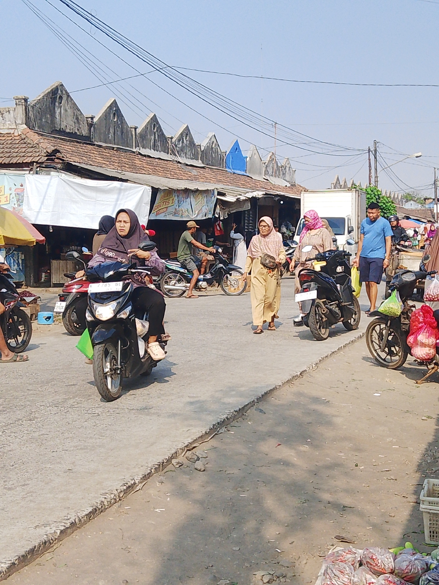 This image captures a street scene in Kalinyamatan, Indonesia, likely a market area, under a bright, clear sky. In the foreground, a woman wearing a hijab and a striped shirt rides a black scooter. She has a purse slung across her body and is wearing light-colored slip-on shoes. Another scooter, red and white, is parked behind her.
To the left, parts of other motorcycles are visible, including a black one with a rider in black. A yellow awning shades a portion of the market stalls in the background.
In the mid-ground, two women walking side-by-side are the central focus. The woman on the left wears a light beige hijab and a patterned dress, with a small bag slung across her shoulder. The woman next to her wears a pink hijab and a similar patterned dress. Behind them, a white truck is parked. A man in a blue t-shirt and dark shorts stands to the right of the truck, holding a yellow shopping bag.
Further to the right, a man wearing a beige hat and green t-shirt is seen on a motorcycle, and other market stalls with various goods, including produce in plastic bags and a white crate, are visible. The architecture of the buildings in the background features pointed, weathered rooflines, characteristic of the region. The ground is paved with concrete and dirt, with some debris present. The overall atmosphere suggests a bustling, everyday market environment.