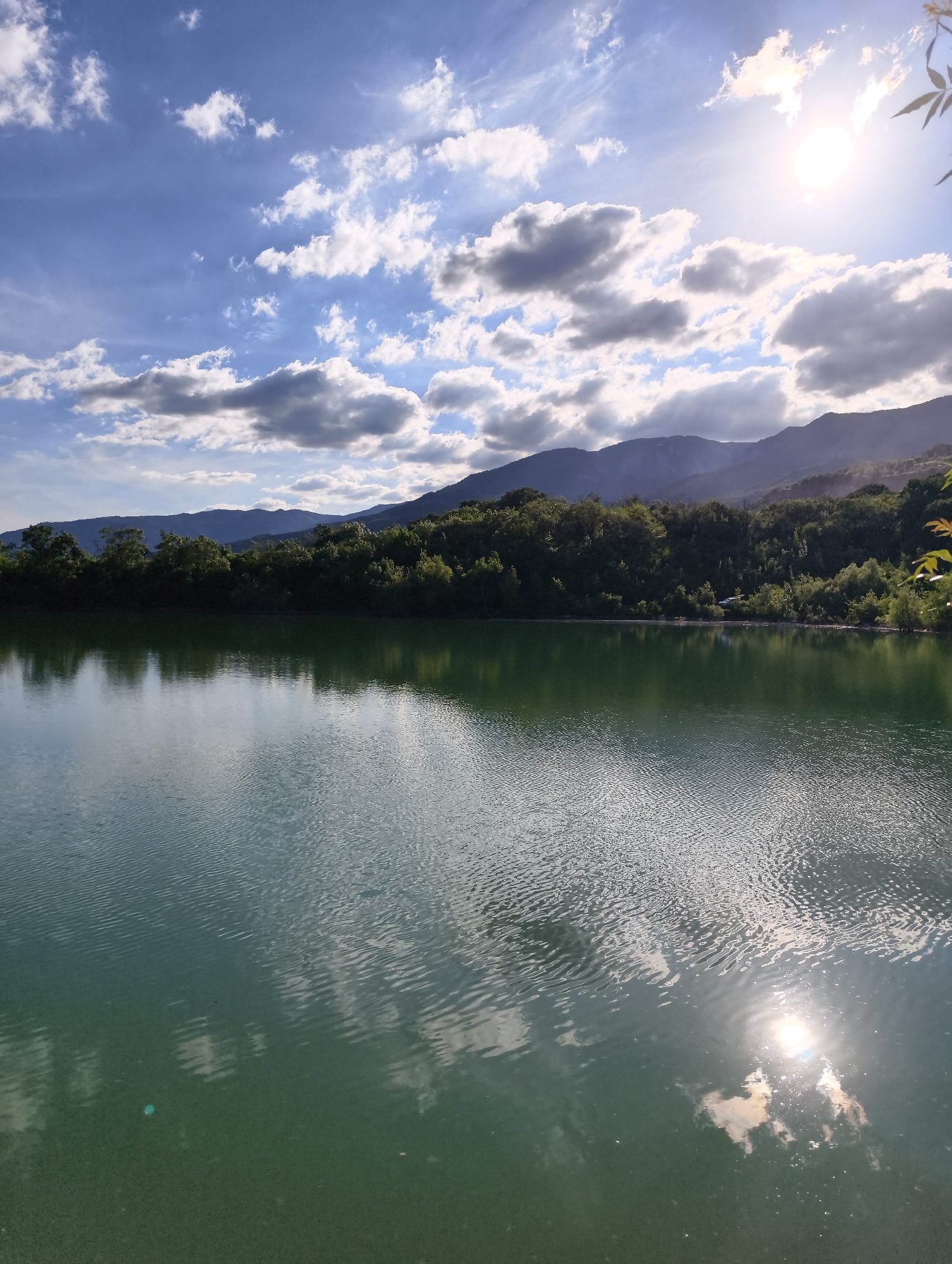 A scenic view captures a tranquil lake in Kiparisnoe, Ukraine. The water, a serene shade of green, reflects the sky and the surrounding landscape. Gentle ripples disturb the surface, creating a mesmerizing pattern of light and shadow. In the background, a dense line of lush green trees forms a vibrant border, separating the water from the distant, hazy mountains. The mountains, layered in shades of blue and purple, recede into the horizon, creating a sense of depth and vastness.
The sky above is a brilliant blue, dotted with fluffy white clouds. The sun, a bright orb in the upper right corner, casts a warm glow, illuminating the scene and creating a shimmering reflection on the water. The presence of sunlight suggests it's daytime, likely late afternoon given the angle of the sun and the soft, diffused light. The overall atmosphere is peaceful and idyllic, evoking a sense of calm and natural beauty. There are no visible people or man-made objects that stand out, emphasizing the untouched, natural character of the location. The image is a still photograph, capturing a moment of serene natural splendor.