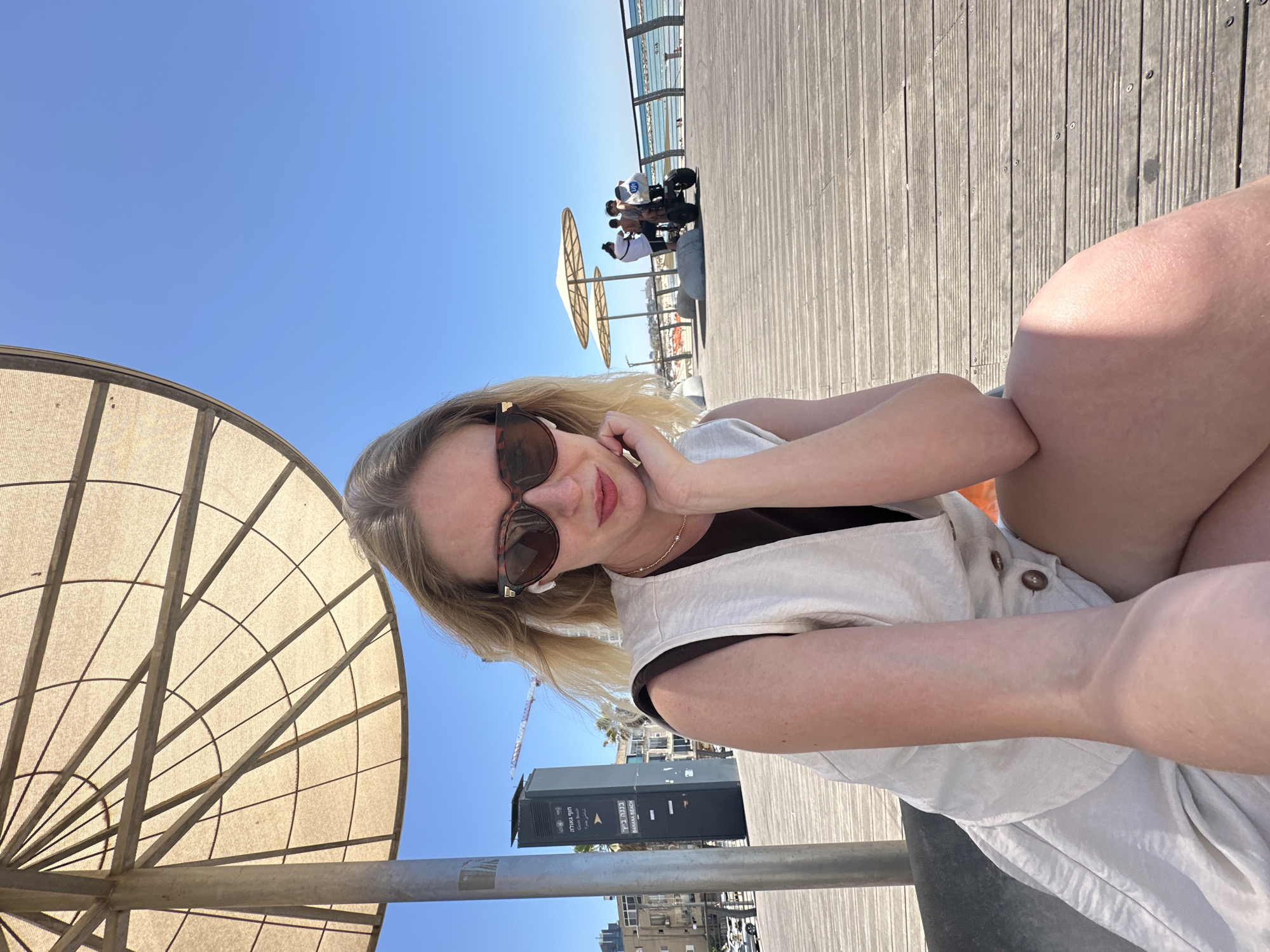 The image depicts a young blonde woman posing for a close-up photo or selfie on a sunny day at a coastal boardwalk. She wears dark sunglasses, a light-colored sleeveless top over a dark shirt, and white wireless earbuds. Her left arm is bent with her hand near her face, and she has a relaxed, slight smile. Her right leg is partially visible in the foreground, suggesting she is seated.
A large, circular, light-colored sunshade, constructed from fabric stretched over a metal frame, dominates the left foreground. To the woman's left, a dark grey utility box or sign is visible. It displays Hebrew text reading "חוף תל-אביב" (Tel Aviv Beach) and "באננה ביץ'" (Banana Beach), along with "BANANA BEACH" in English.
In the background, a wooden boardwalk stretches to the right and into the distance. Several indistinct figures are seen strolling or standing, one possibly on a scooter. Further back, another smaller sunshade appears. Beyond the boardwalk, a strip of the blue sea and a distant cityscape with buildings, palm trees, and a construction crane are visible under a clear blue sky. The bright sunlight indicates a mid-day scene, conveying a relaxed and pleasant atmosphere on a beach promenade in Tel-Aviv, Israel.