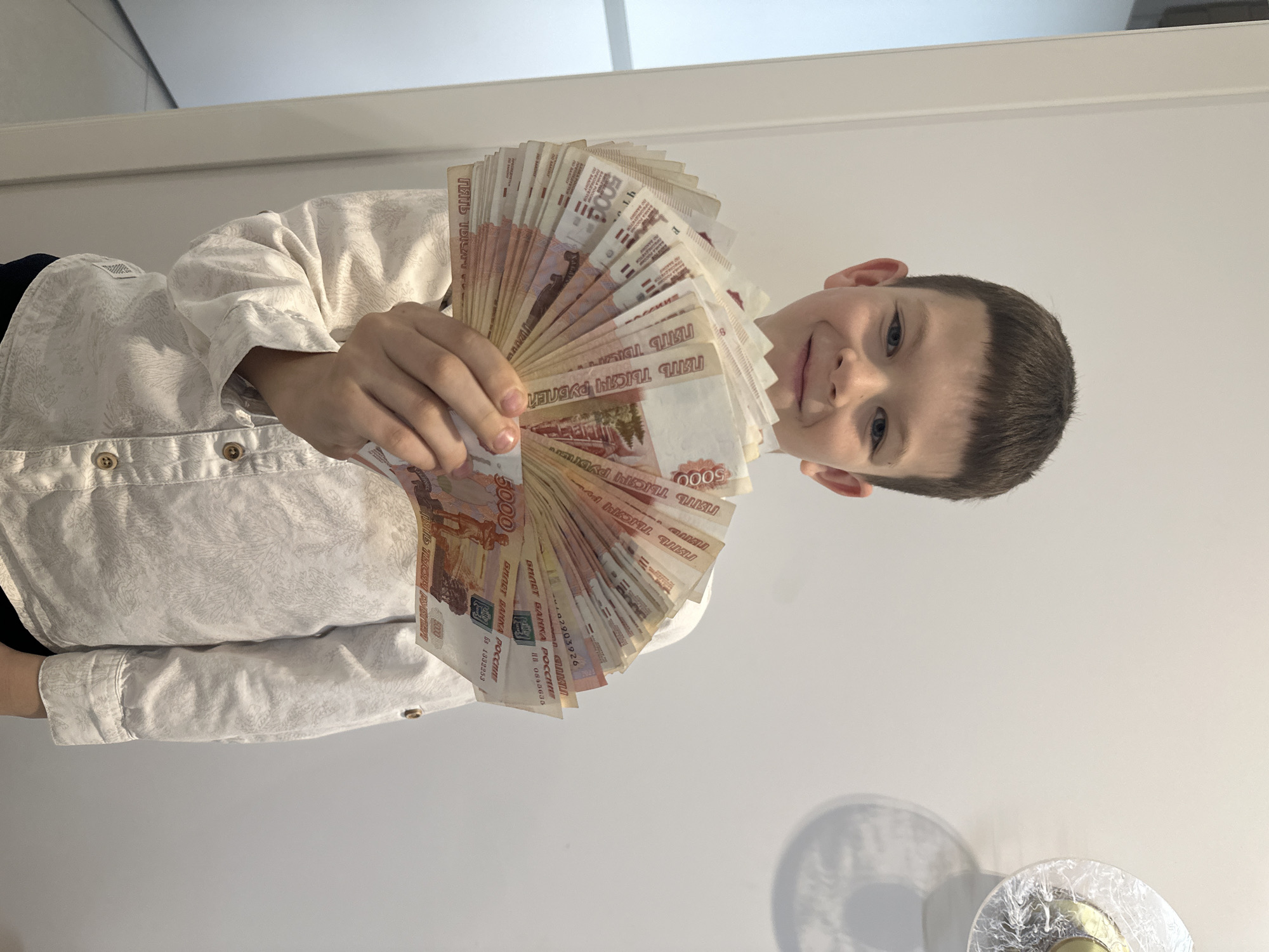A young boy with brown hair is holding a large fan of 5000 Russian Ruble bills. He is wearing a light-colored, button-down shirt. The background is a plain white wall with what appears to be the edge of a cabinet or door frame. There is also a decorative wall light fixture in the lower right corner of the image.