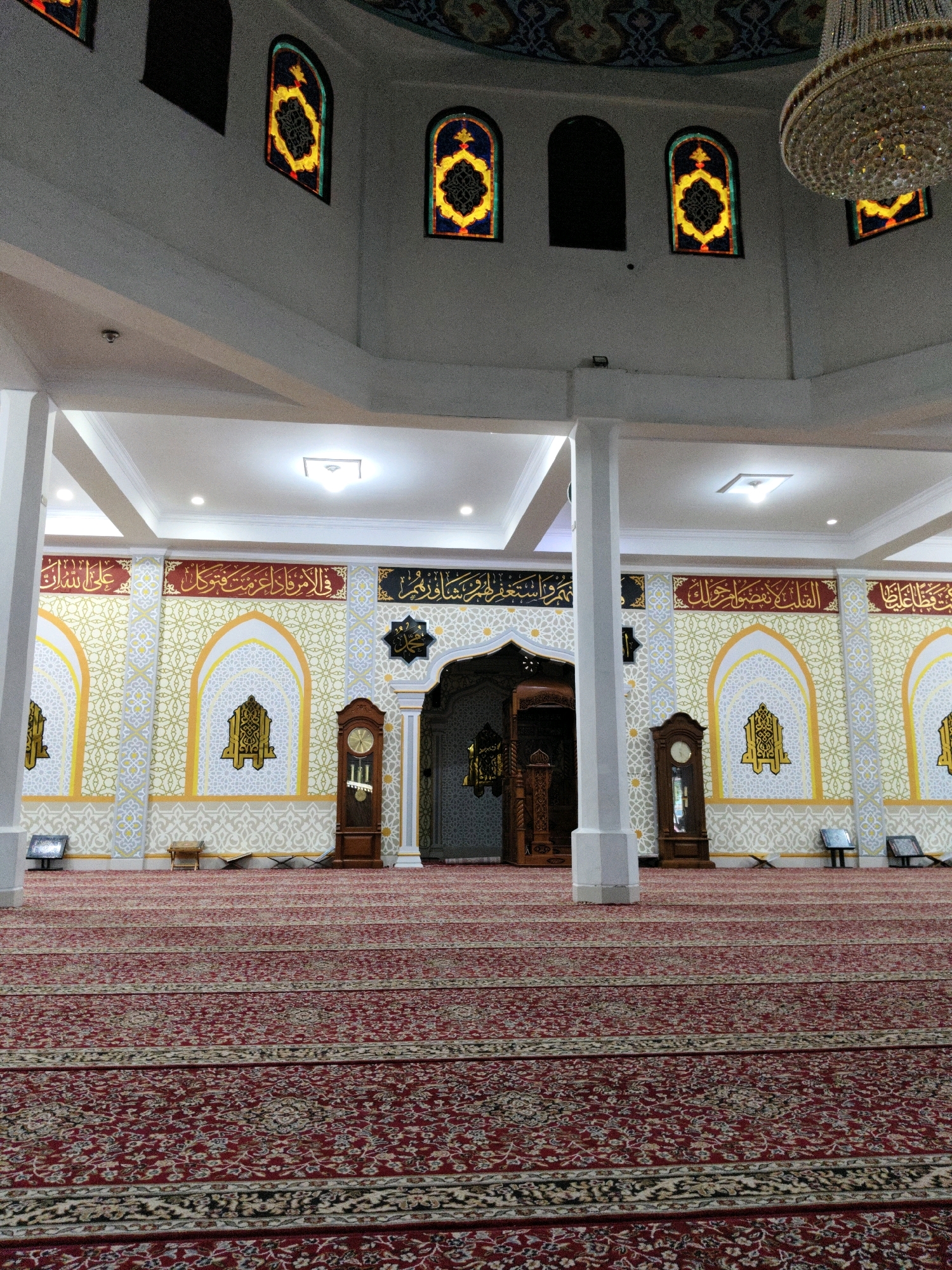 The image shows the interior of the Islamic Center Lhokseumawe, a mosque located in Lhokseumawe, Indonesia. The foreground is dominated by a large, intricately patterned red carpet with cream and black stripes running horizontally. In the midground, white pillars support the ceiling. The walls are decorated with a repeating geometric pattern in cream and gold, interspersed with arched niches containing calligraphy and ornate designs. Two tall wooden grandfather clocks stand against the walls.
To the left, a wooden lectern is visible. Further back, a grand wooden entrance with intricate carvings leads into another part of the building. Above the walls, stained-glass windows with colorful geometric designs are set into the upper walls, which are painted white. A large, crystal chandelier hangs from the ceiling, partially visible on the right side of the frame. The overall impression is one of grandeur and intricate Islamic artistry within a mosque setting.
