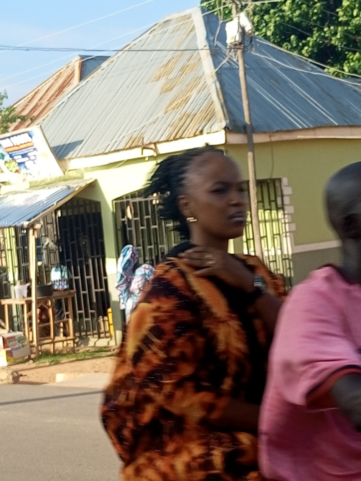 In Tempure, Nigeria, a woman with braided hair is captured in profile, wearing a patterned orange and black dress and a wristwatch. She stands on a paved road next to a light green building with barred windows, facing a building with a corrugated metal roof. To her right, another person in a pink t-shirt is partially visible. The background reveals a street scene with a utility pole, trees, and various structures.