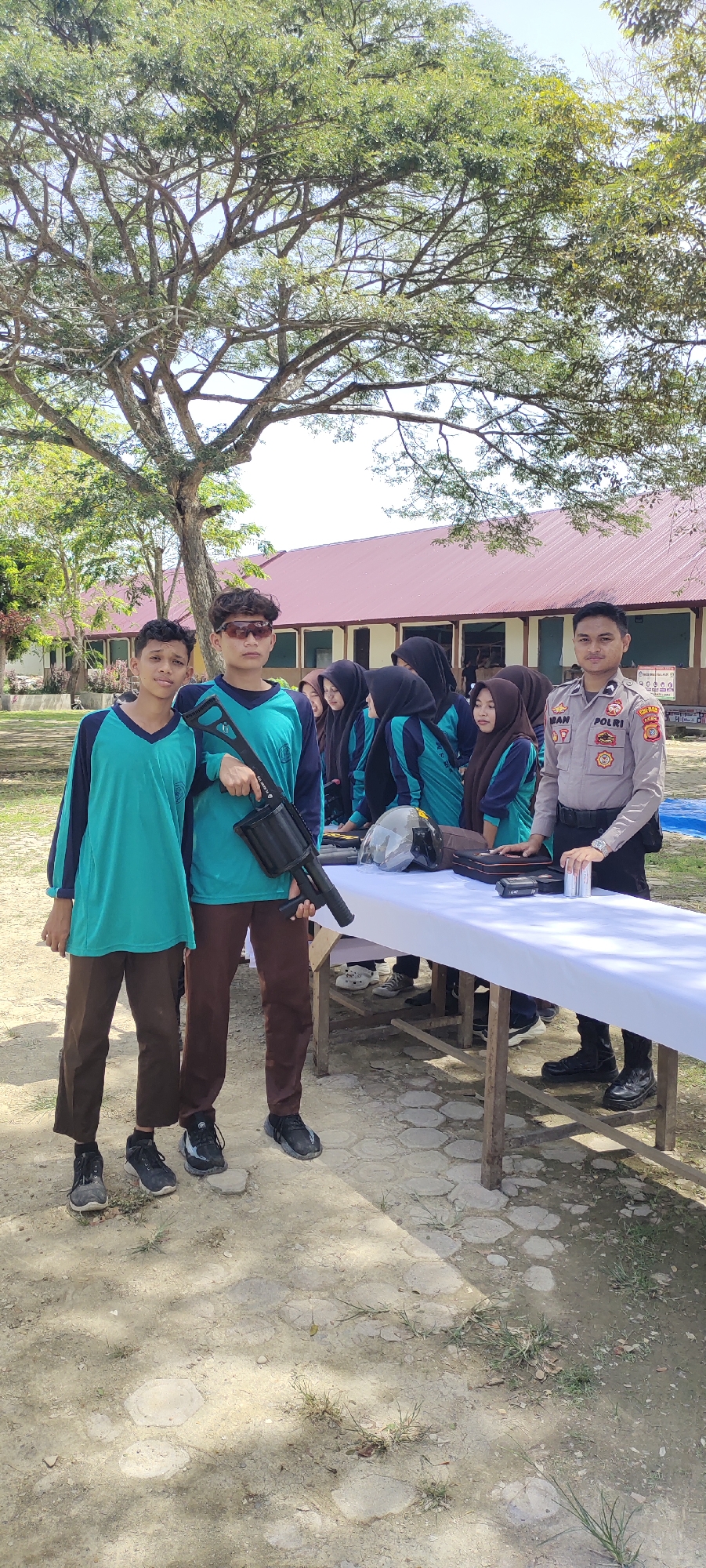 The image shows a group of people outdoors, likely in a schoolyard, as there is a building with a red roof and multiple windows visible in the background. Two young males dressed in matching turquoise and dark brown uniforms stand in the foreground. One of them is holding what appears to be a black prop gun. A white table, covered with a white tablecloth, is set up nearby. Behind the table, there is a group of young females wearing headscarves, along with a male police officer dressed in a uniform. The ground is paved with light-colored stones. A large tree stands in the background. The location may be Langnibong, Indonesia.
