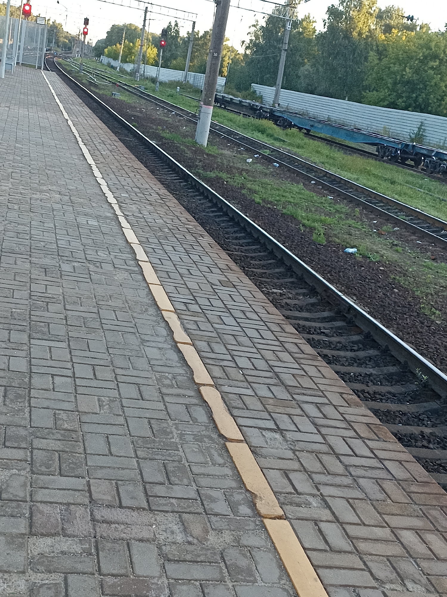 The image depicts a train station platform in Chekhov, Russia. The platform is paved with grey rectangular bricks arranged in a herringbone pattern, with a yellow stripe along the edge next to the train tracks. The train tracks themselves are visible, with ballast and green grass growing between them. In the distance, there are trees and a metal fence. A blue train car is visible on the tracks. There are also traffic lights visible, which are currently red. The sky is a bright, hazy blue. No people are visible in the image.