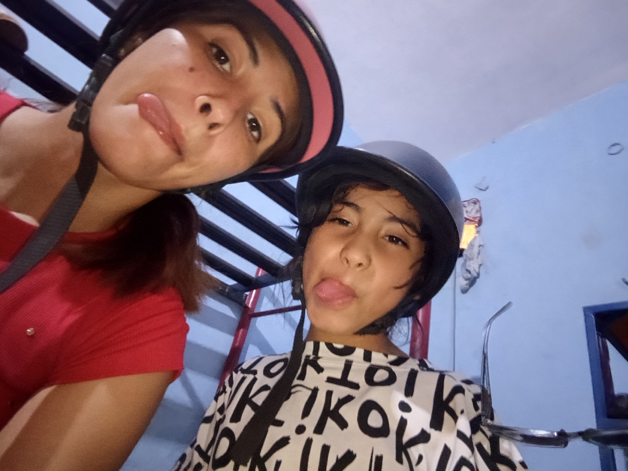 Two individuals are posing for a selfie indoors. The person on the left, wearing a red short-sleeved shirt and a pink-trimmed black helmet, is sticking out their tongue. The person on the right, wearing a black and white patterned shirt and a plain black helmet, is also sticking out their tongue. A set of stairs with red and white accents is visible in the background. The wall behind them is light blue, and a partial view of what appears to be a doorway or window frame can be seen on the right. This image provides no context about Juan Griego.