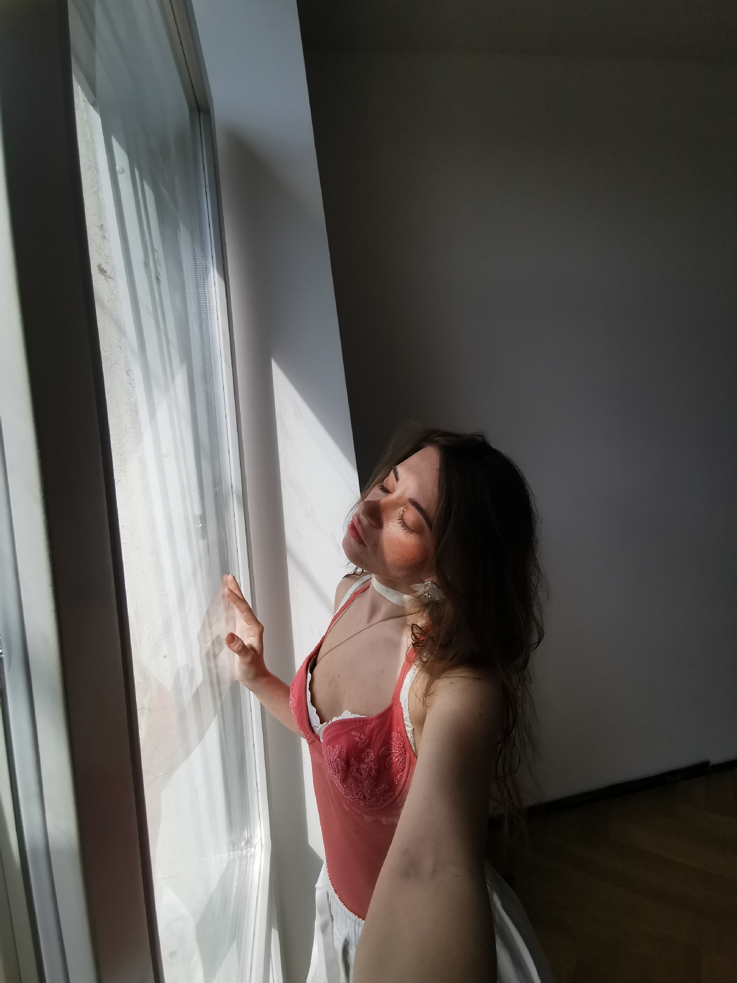 A woman with light skin and brown hair stands in front of a window with white curtains. She is wearing a coral-colored lingerie top with white lace and a white collar necklace. Sunlight streams through the window, illuminating her face and casting shadows across the room. The background consists of a white wall and a dark grey wall, and the floor is made of wood. The location is a room in Moscow, Russia.