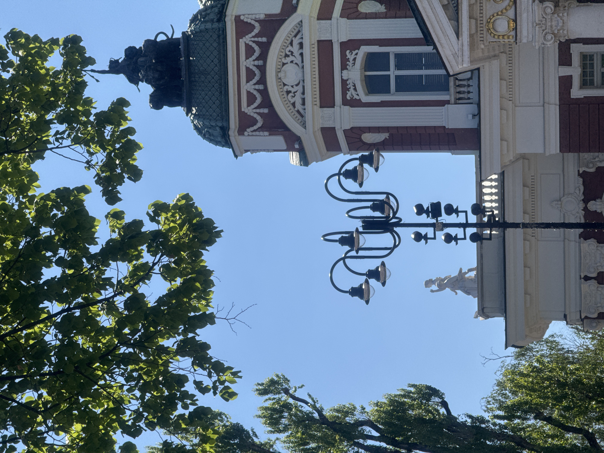 The image shows a view of a building in Sofia, Bulgaria, from a low angle, looking upwards. The building has a red and white facade with ornate details, including a tower with a domed top. There is a statue on the roof of the building. A black lamppost with multiple lamps is positioned in front of the building. Green trees with leaves surround the building and lamppost. The sky is blue and clear.