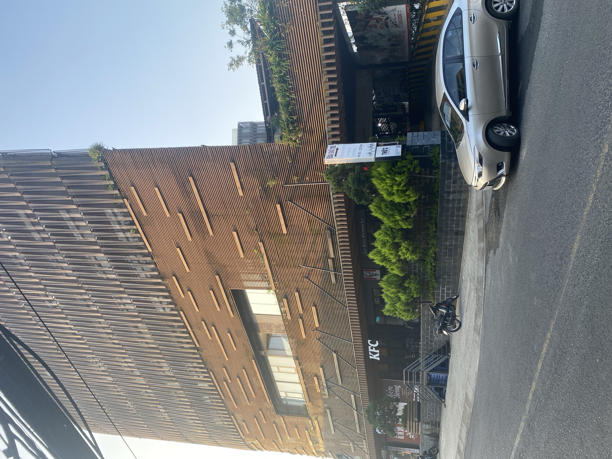The image shows the KFC restaurant in Da Lat, Vietnam. It is located in a modern building with a facade of horizontal wooden slats. The KFC logo is visible on the front of the building. A light-colored car is parked on the side of the road. The environment appears to be urban. A sign in Vietnamese is visible near the entrance, and some greenery is present. A motorcycle is parked on the sidewalk. The weather seems clear and sunny.