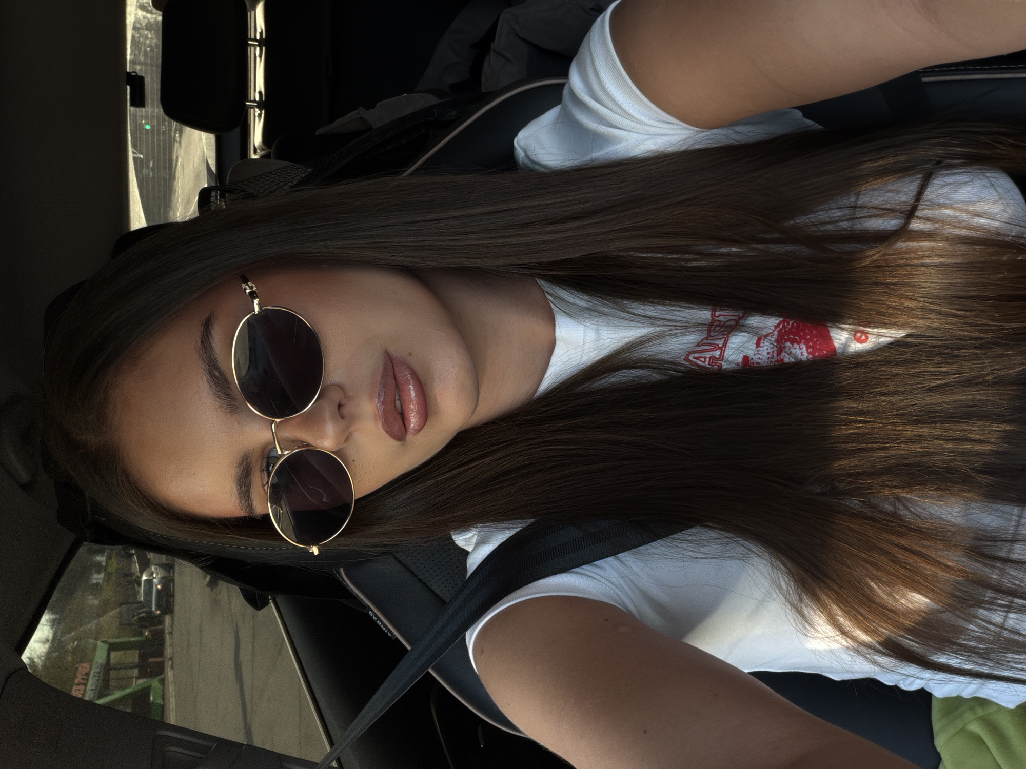 A woman with long brown hair, sunglasses, and a white t-shirt is sitting in a car. She is wearing a seatbelt. The location appears to be Krasnoyarsk, Russia, as evidenced by a sign in the background visible through the car window written in Russian.
