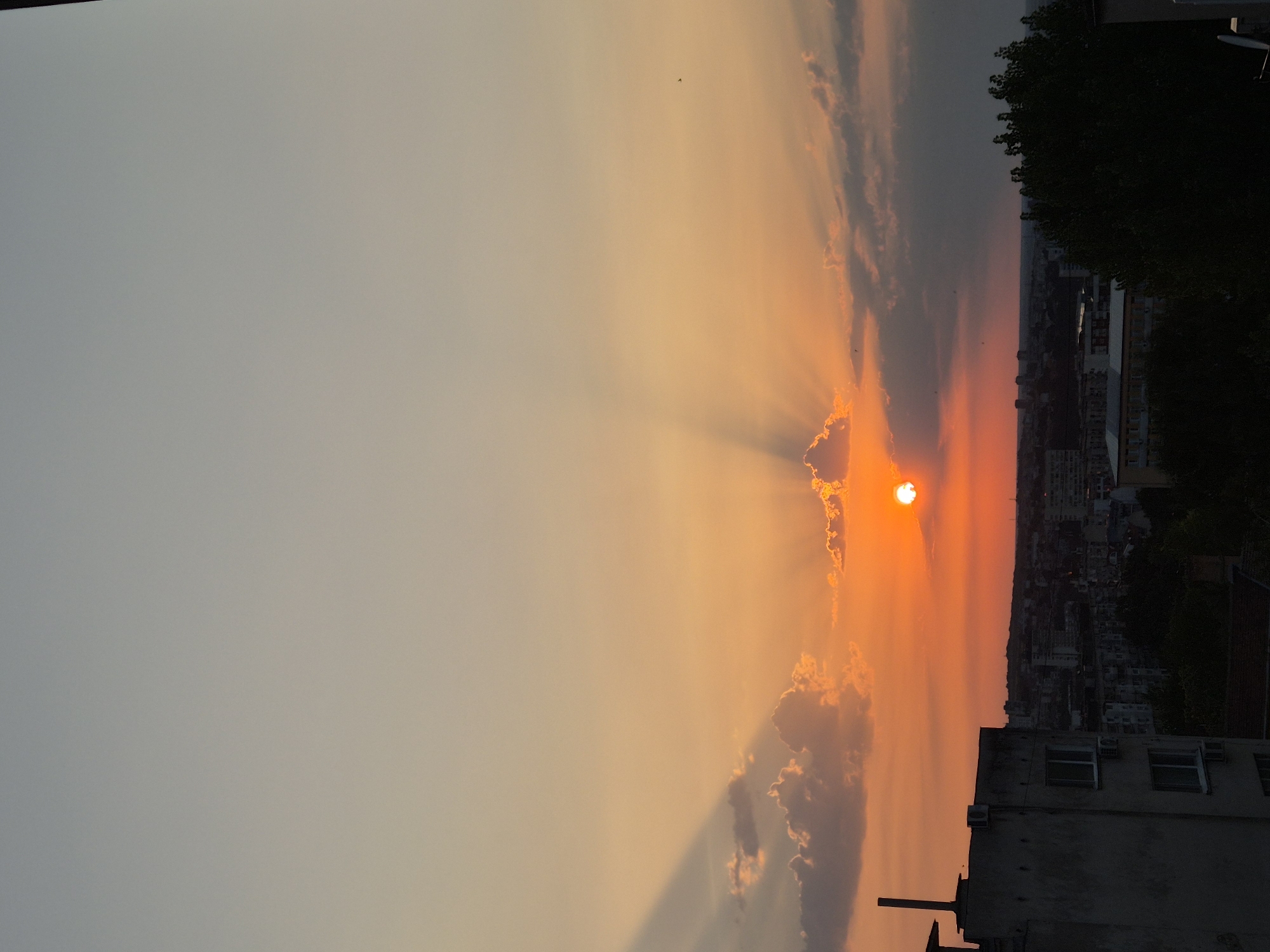 The image captures a vibrant sunset over a city. The sun, a bright orange orb, is partially obscured by clouds, casting dramatic rays of light across the sky. The sky transitions from a pale, dusky gray at the top to warm oranges and reds closer to the horizon. Silhouetted buildings and trees form a dark, dense cityscape in the foreground and middle ground. A portion of a building, appearing to be under construction or in disrepair, is visible on the left side, with a pipe-like structure extending upwards from its roof. The overall scene is one of a city bathed in the warm glow of the setting sun, with a mix of urban structures and natural elements.