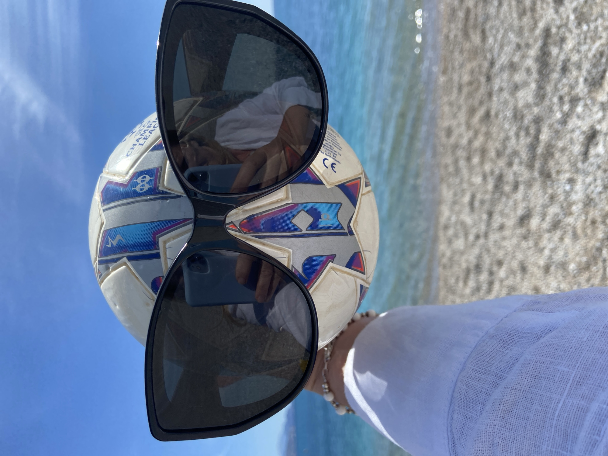 The image shows a person holding a white soccer ball with blue and silver designs, in front of a beach scene. The person is wearing a white shirt or blouse, and a bracelet of some sort. Sunglasses are positioned on the soccer ball, reflecting the person holding the ball and their smartphone. The beach is in the background, with blue water meeting a sandy or pebbly shore. The location is Prijevor, Montenegro. The sky is blue and clear.