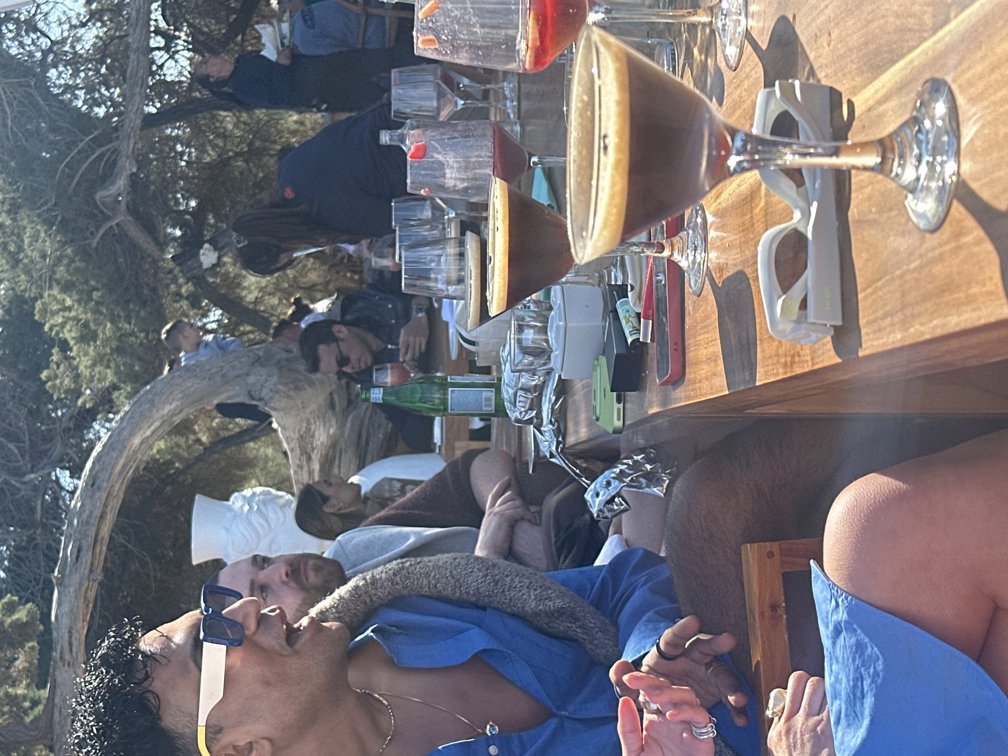 A daytime scene in Sant Josep de sa Talaia, Spain shows several people gathered around a wooden table with drinks. On the table are cocktails in martini glasses, some partially consumed, and other glassware, a water bottle, sunglasses, and personal items such as a phone. The people are dressed casually and appear to be enjoying a social gathering. In the foreground, a person wearing sunglasses and a blue shirt is visible, with another person sitting beside them. Behind the table, other people are seated or standing, some of whom are in conversation. A white, classical-style bust and a curved tree trunk are visible in the background, suggesting an outdoor setting.