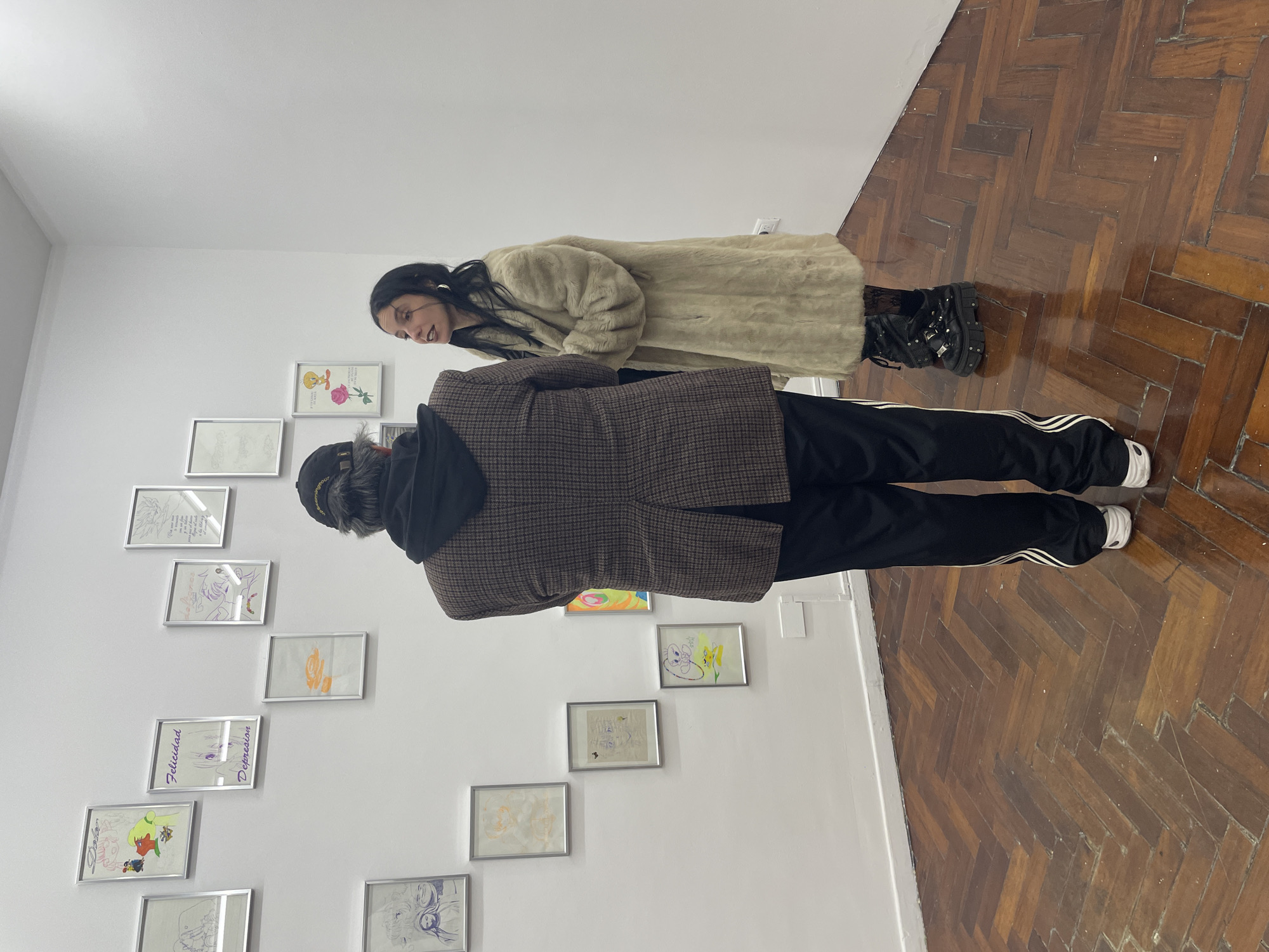 In a brightly lit gallery setting with parquet flooring, two individuals are observed viewing artwork on a white wall. The person closest to the camera wears a brown checkered blazer over a black hooded sweatshirt, with black Adidas track pants featuring white stripes and white sneakers. The other individual, positioned further into the room, is wearing a long, light-colored fur coat and black boots. The artwork consists of framed drawings, several of which are visible and contain images, colors, and text such as "Felicidad" and "Depression." The location is inferred to be in Buenos Aires, Argentina, due to the mention in the prompt.